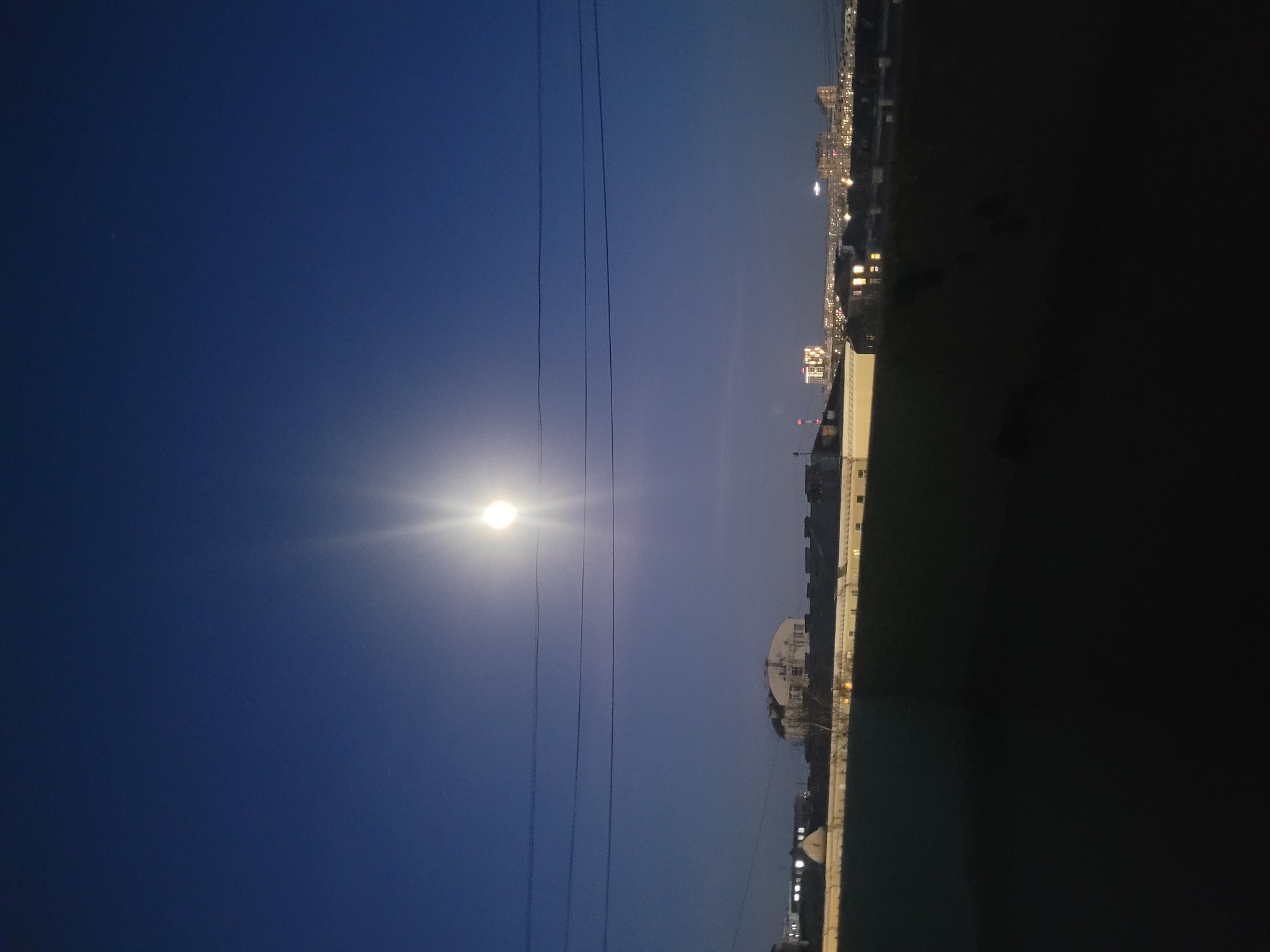 The image shows a nighttime scene in Izhevsk, Russia. The moon, a bright orb, is visible in the sky and has power lines running in front of it. Below the power lines is an urban landscape with buildings and lights. The right side of the image is dark, but buildings are visible. Some lights are visible on the tops of the buildings on the right side of the image.
