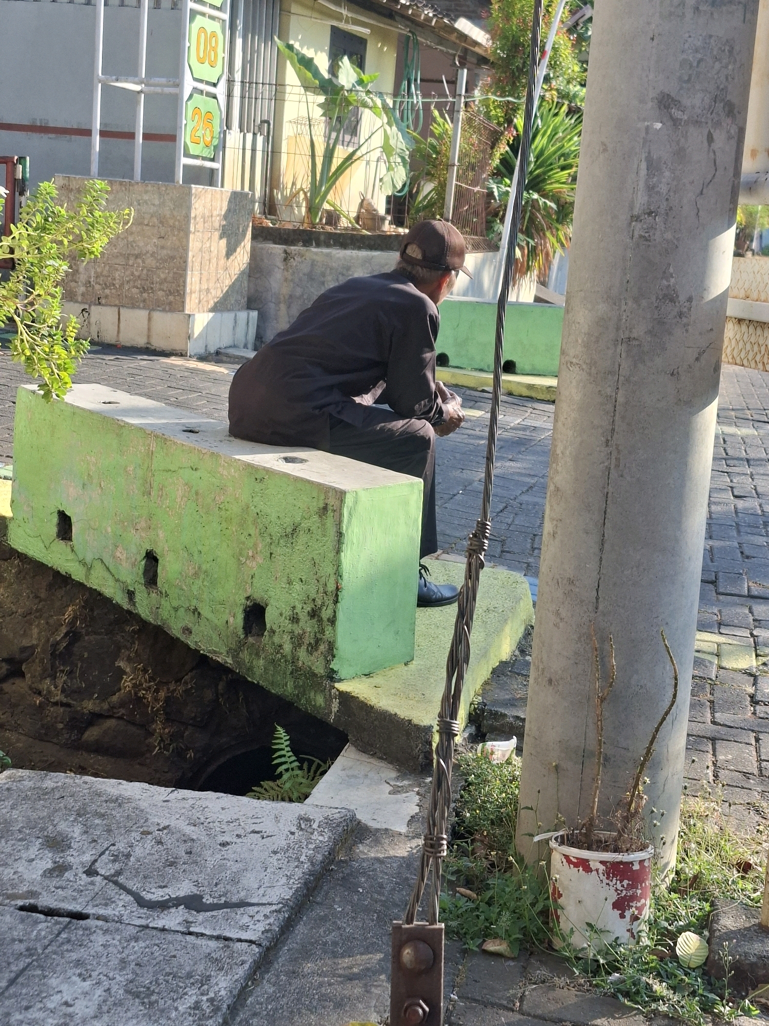 The image depicts a man wearing a dark shirt and cap, sitting on a green concrete structure. He is facing away from the camera, looking towards a hole in the ground next to the structure. The man is holding a phone in his hands. The scene appears to be outdoors, possibly in a public area, with a cobblestone pavement visible. A large concrete pole with wires attached stands to the right of the man. In the background, there are buildings and lush greenery. The overall impression is one of a quiet, possibly reflective moment in an urban or semi-urban setting. The specific location in Semarang, Indonesia, is not identifiable from the visual elements alone.