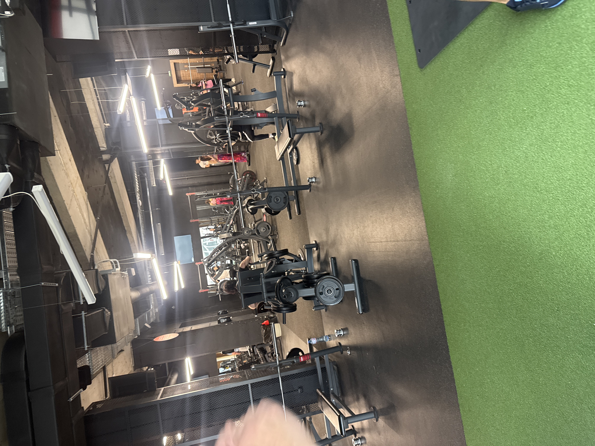 The image shows the interior of a gym in Unknown, Unknown. The scene features numerous weightlifting machines and equipment arranged on a black floor. Some individuals are using the equipment. Overhead, there are several fluorescent tube lights and various industrial-style fixtures. Part of the floor on the right side is covered with artificial green turf.