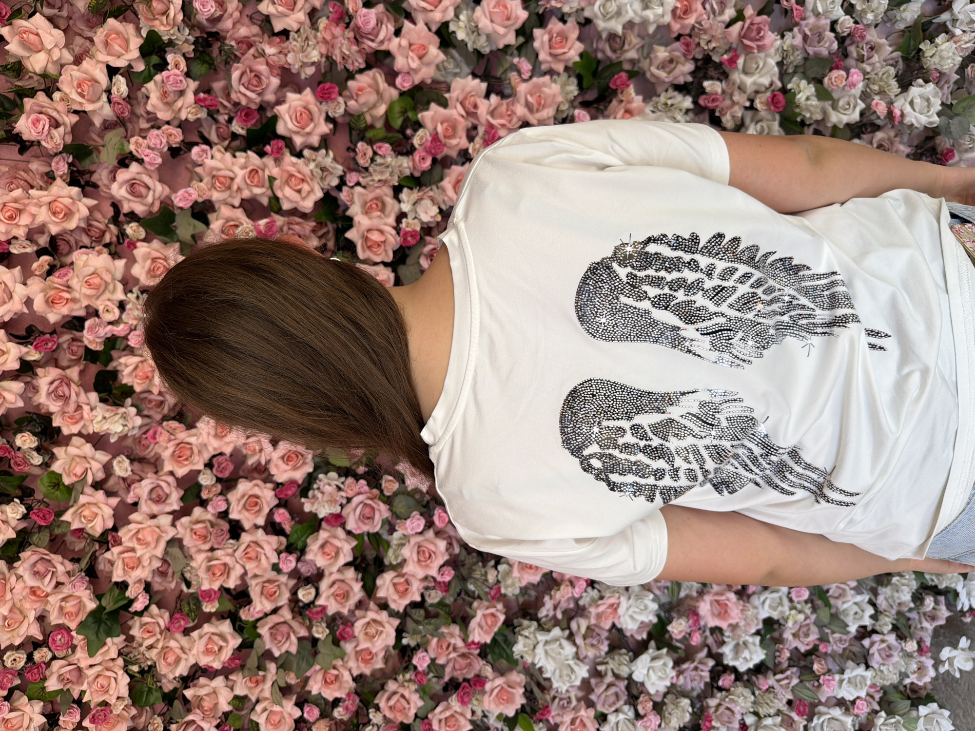 The image depicts a person from the back, wearing a white t-shirt adorned with a rhinestone wing design on its back. The person has brown hair and is standing in front of a wall covered in a dense arrangement of pink and white artificial roses. The setting appears to be indoors or a covered outdoor space, given the artificial nature of the floral display. The lighting is bright and even, suggesting it could be studio lighting or natural light from a source not visible in the frame. The overall aesthetic is feminine and decorative. There is no information or visual evidence within the image to determine its location in the city of Ternopil, Ukraine.