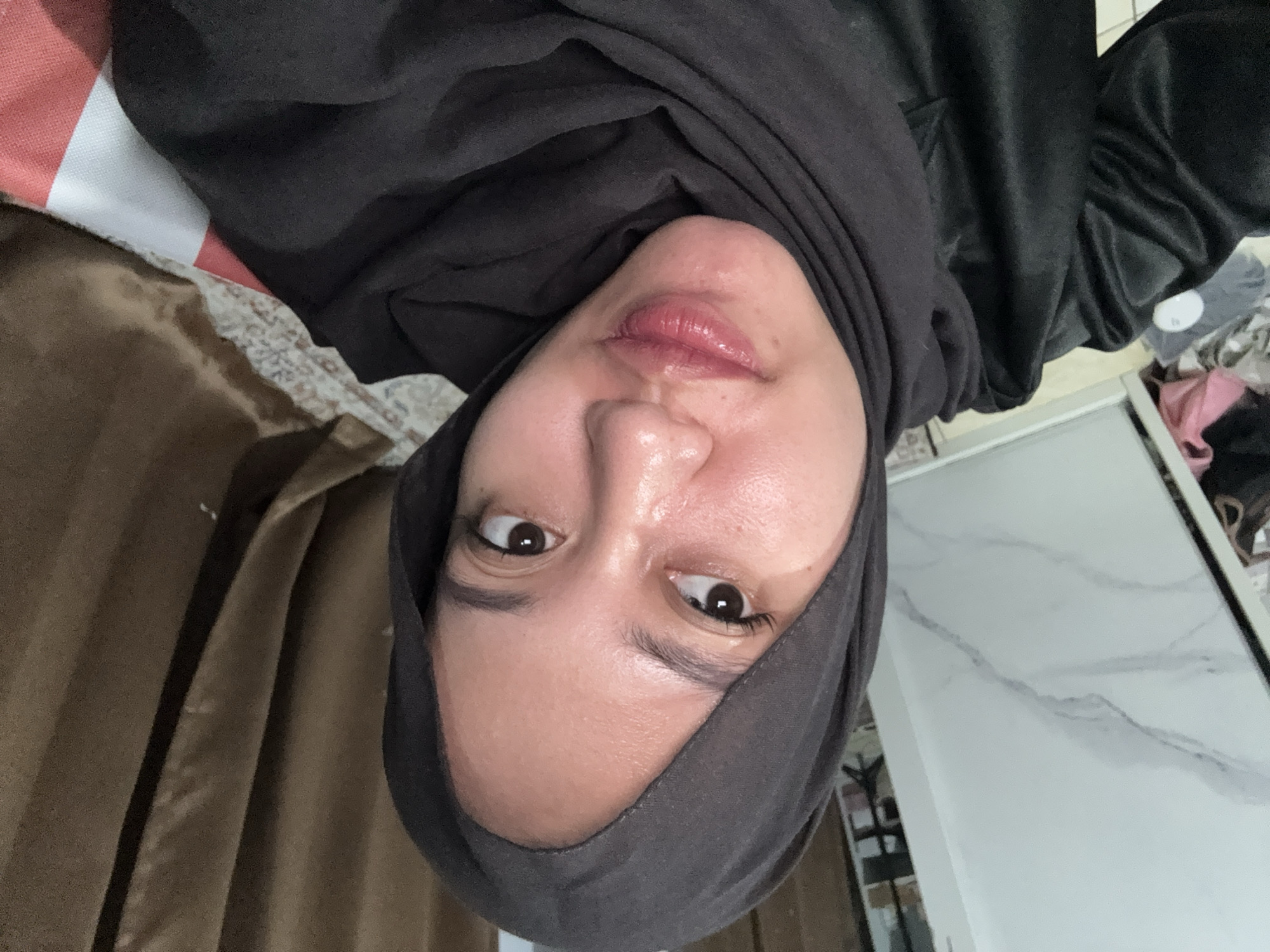 A young woman wearing a dark grey hijab and a black top is looking to her left. She has makeup on, including mascara and lipstick. Her skin appears slightly shiny. The background shows a wall with marble-like patterns on the left, brown curtains on the right, and a patterned rug on the floor. There are also some clothing items and a white and red striped object visible. The lighting in the image is soft.
