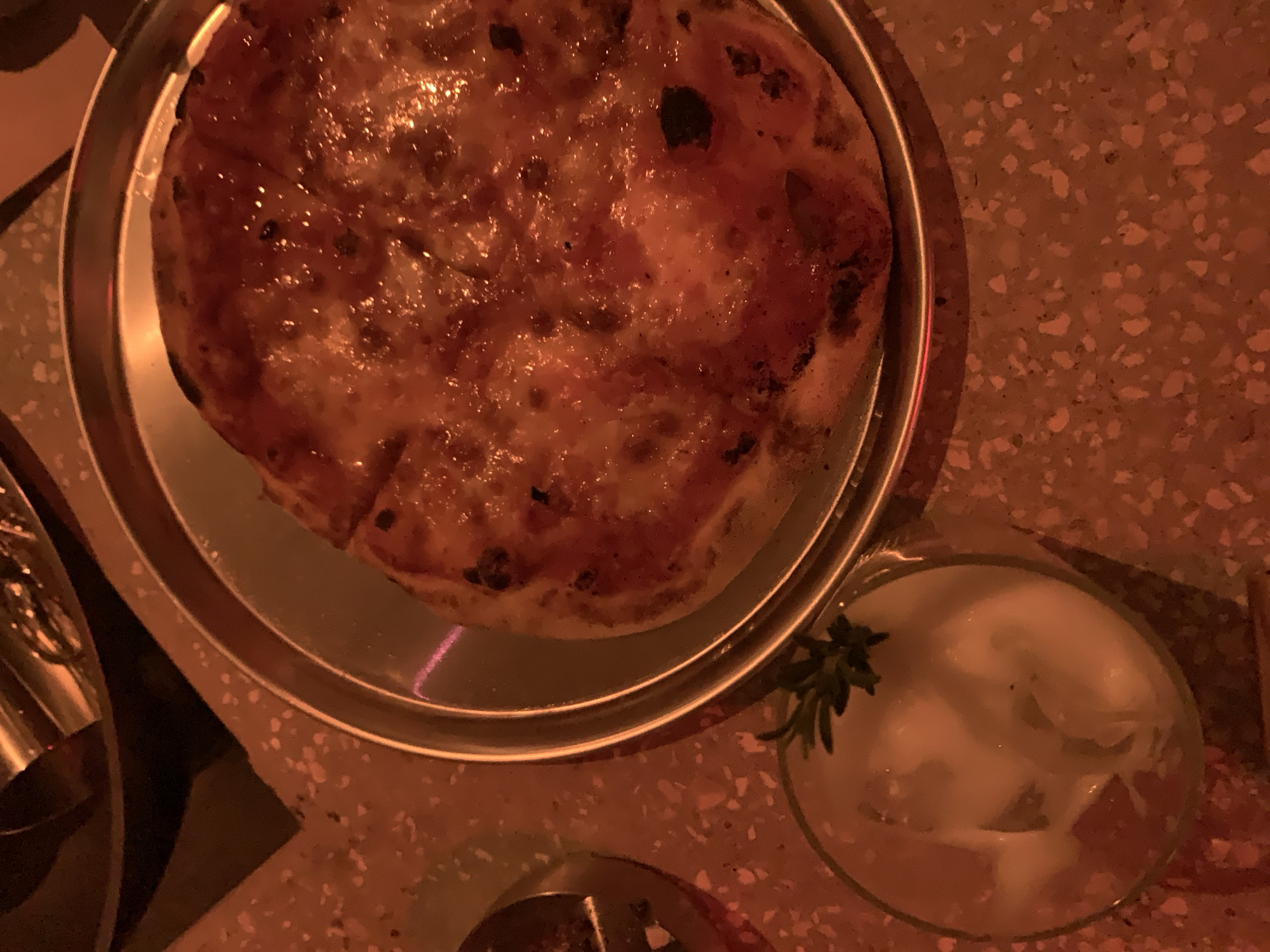 A top-down, close-up shot shows a whole pizza on a metallic plate, next to a small glass filled with a white dip and a sprig of green herb. The pizza is golden brown with some darker charred spots, suggesting it's freshly baked. The plate is round and silver, with a slightly raised rim. The glass is clear and appears to contain a creamy substance with some thinly sliced white pieces and a small green herb garnish on top. The background is a textured surface, possibly a table, with a speckled pattern in shades of orange and brown. The lighting is dim and warm, casting an orange hue over the entire scene, suggesting an indoor setting, possibly a restaurant in Tel Aviv, Israel. There are no people or any interaction visible in the image.