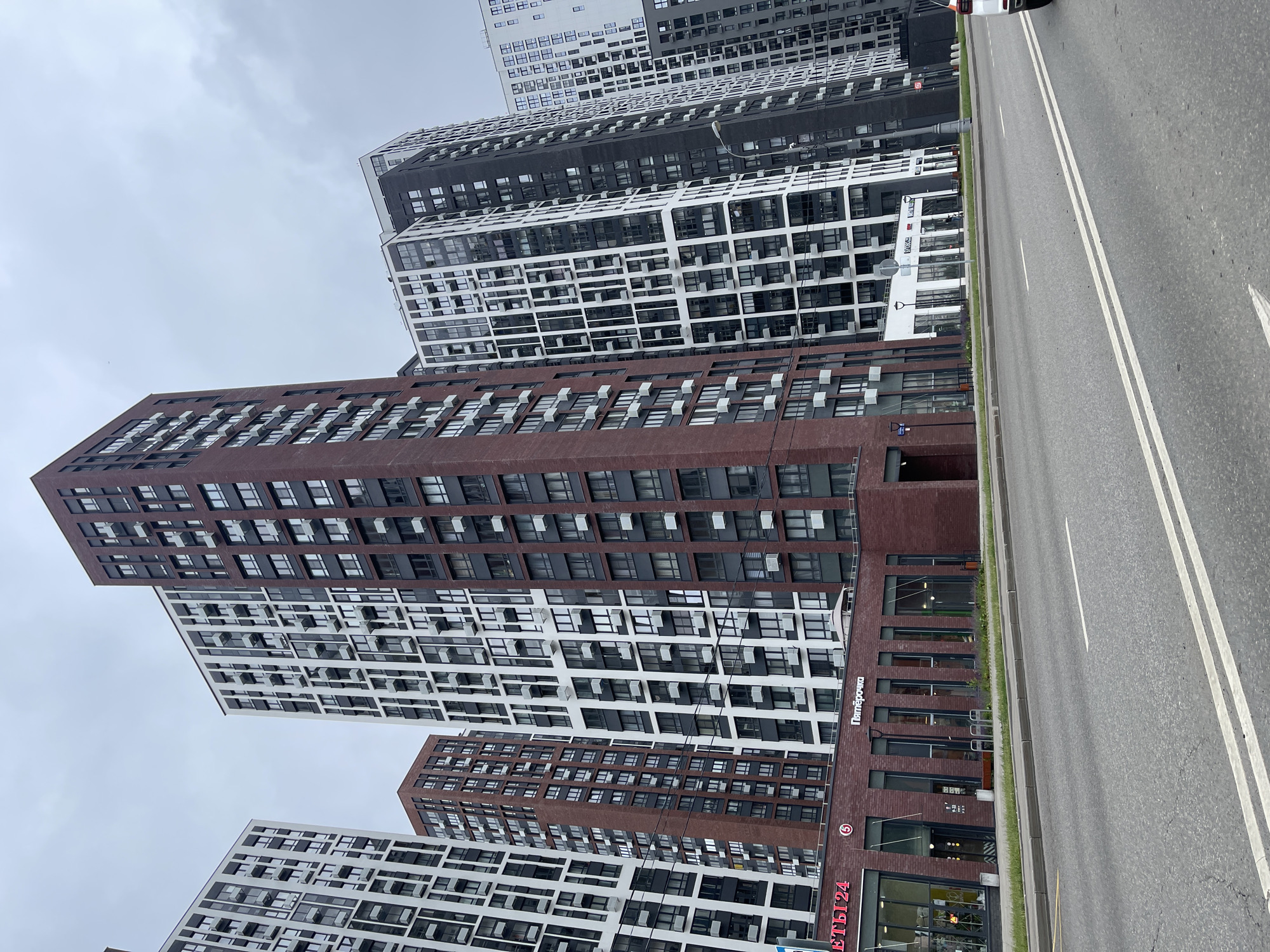 The image shows a modern residential complex with several high-rise buildings lining a wide street in Moscow, Russia. The buildings are contemporary in design, featuring a mix of dark red brick and white and black facades with numerous windows. Some buildings have balconies with air conditioning units visible. The street in the foreground is paved and has double white lines marking traffic lanes. There are sidewalks and some greenery along the buildings. The sky is overcast. Signs are visible on the lower levels of some buildings, including one in Russian that appears to be for a store or business. The scene depicts an urban residential area on a cloudy day.