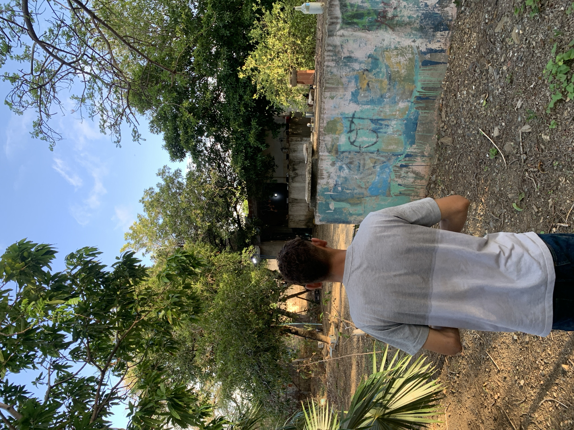 A young man with short, curly brown hair stands with his back to the camera, looking towards an outdoor area in Santa Ana, Venezuela. He is wearing a two-toned grey t-shirt and blue jeans. The environment appears to be a somewhat neglected yard or lot, with dry soil, scattered rocks, and debris. Lush green trees with dense foliage are visible in the background, casting shadows and creating a dappled light effect. In the mid-ground, a weathered concrete structure, possibly a wall or building remnant, is adorned with colorful, abstract graffiti, including drips of blue and green paint. Several objects are present on or near this structure, including what appears to be a light fixture, some pipes, and a white plastic bottle. The sky above is a clear blue with a few wispy clouds. The overall impression is of a rural or semi-rural setting within the city, characterized by a blend of natural elements and signs of human habitation and decay.