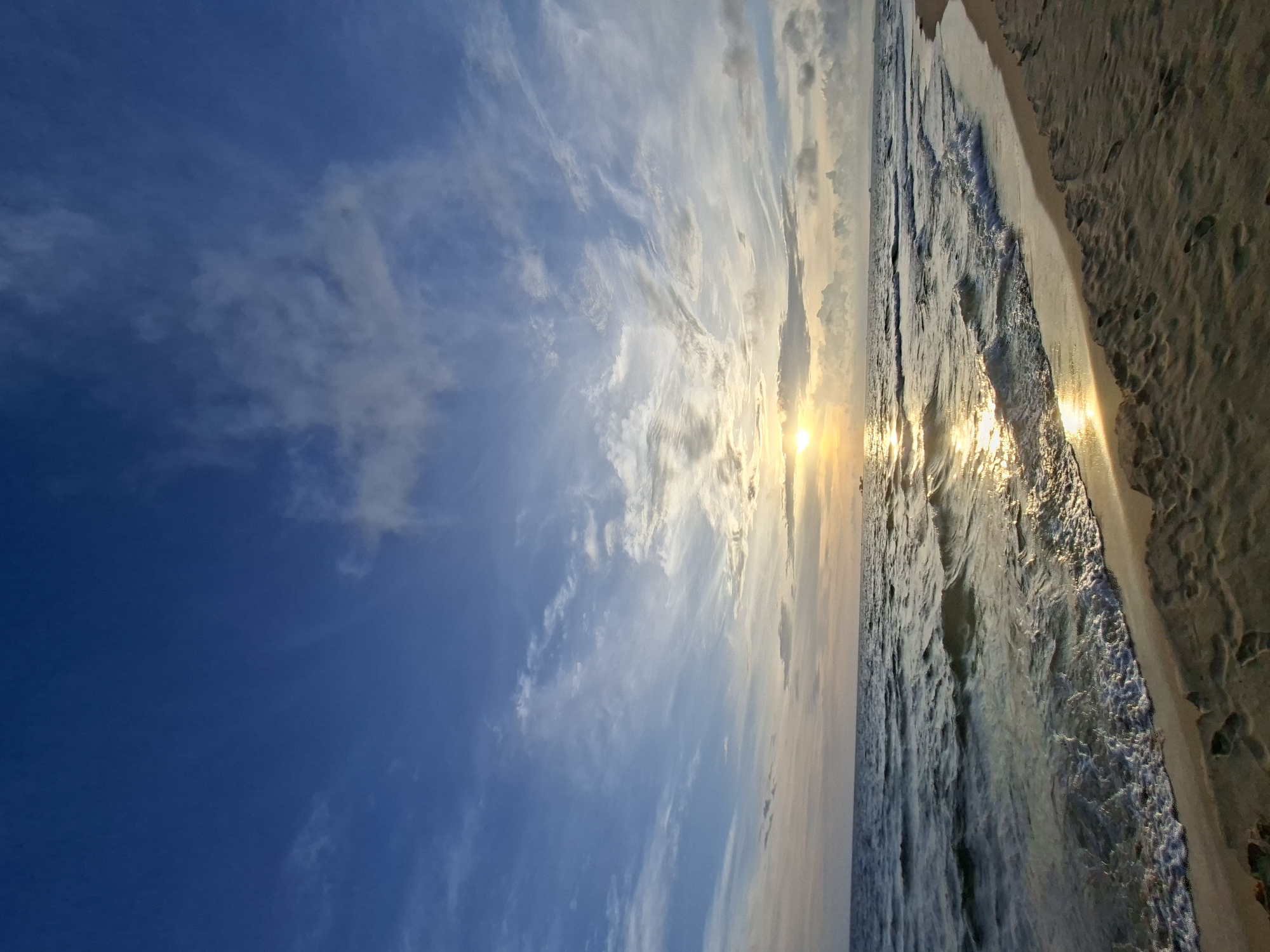 The image depicts a tranquil beach scene at sunset or sunrise. The sky is a vibrant blue with wispy clouds scattered across it, some catching the golden light of the sun which is partially obscured by clouds near the horizon. The sun's reflection shimmers on the surface of the ocean, creating a path of light that stretches towards the shore. Gentle waves roll onto a sandy beach, leaving a foamy edge where they meet the sand. The sand itself shows faint footprints, suggesting recent activity. The overall impression is one of peace and natural beauty, with the warm hues of the sky and water creating a serene atmosphere.