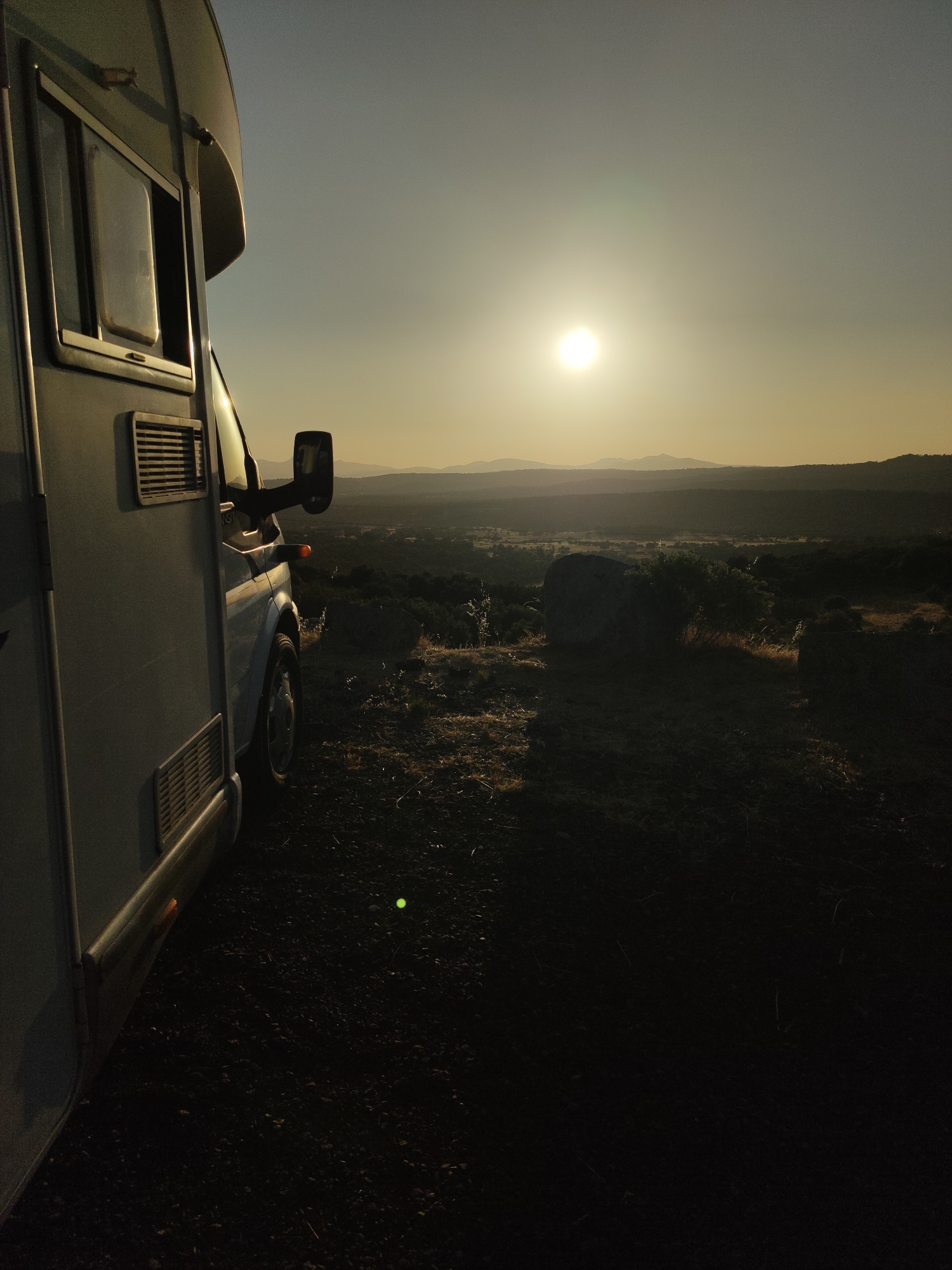 An exterior, low-angle, right-facing shot captures a side view of a light-colored camper van parked on a dirt and rocky surface at dusk. The sun, a bright orb in a hazy sky, is positioned high and slightly to the left of the frame, casting a warm, golden light that illuminates the scene. The camper van occupies the left portion of the image, its window and exterior vents visible. The right side of the image opens to a vast landscape, characterized by rolling hills and mountains in the distance, receding into soft focus. The foreground consists of rough terrain with scattered rocks and dry vegetation, some of which are illuminated by the setting sun. A large, rounded boulder is prominent in the middle ground to the right, partially obscured by a bushy plant. The overall atmosphere is serene and contemplative, suggesting a moment of peaceful repose in a remote natural setting. This scene is located in the municipality of Ledrada, Salamanca, Spain.