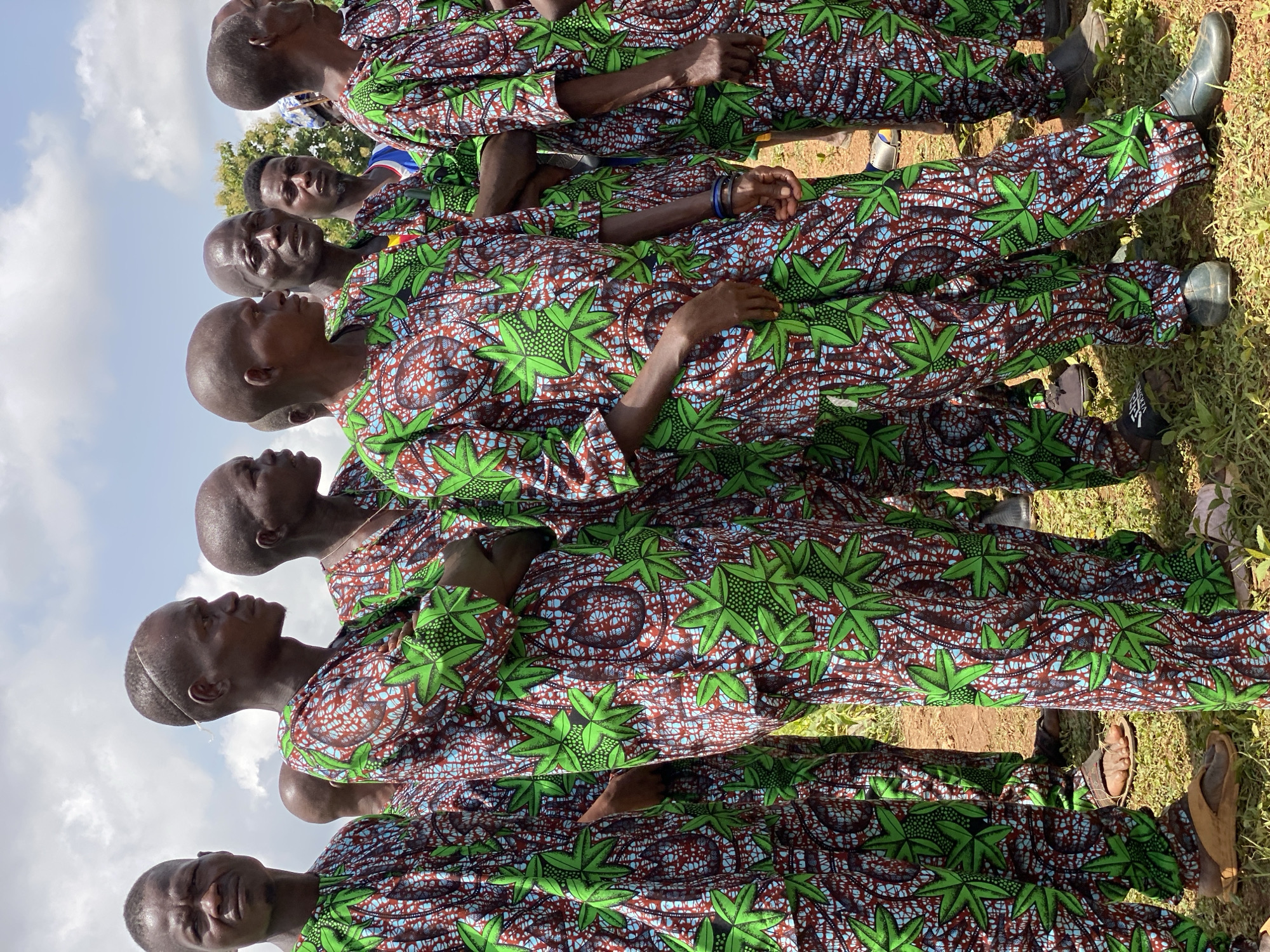 A ground-level, outdoor shot shows a line of men wearing the same patterned long-sleeved shirt and pants standing in what appears to be a grassy area. The sky is visible above, with some clouds present. The men are looking toward the same direction. Maigizo, Nigeria, is not confirmed.