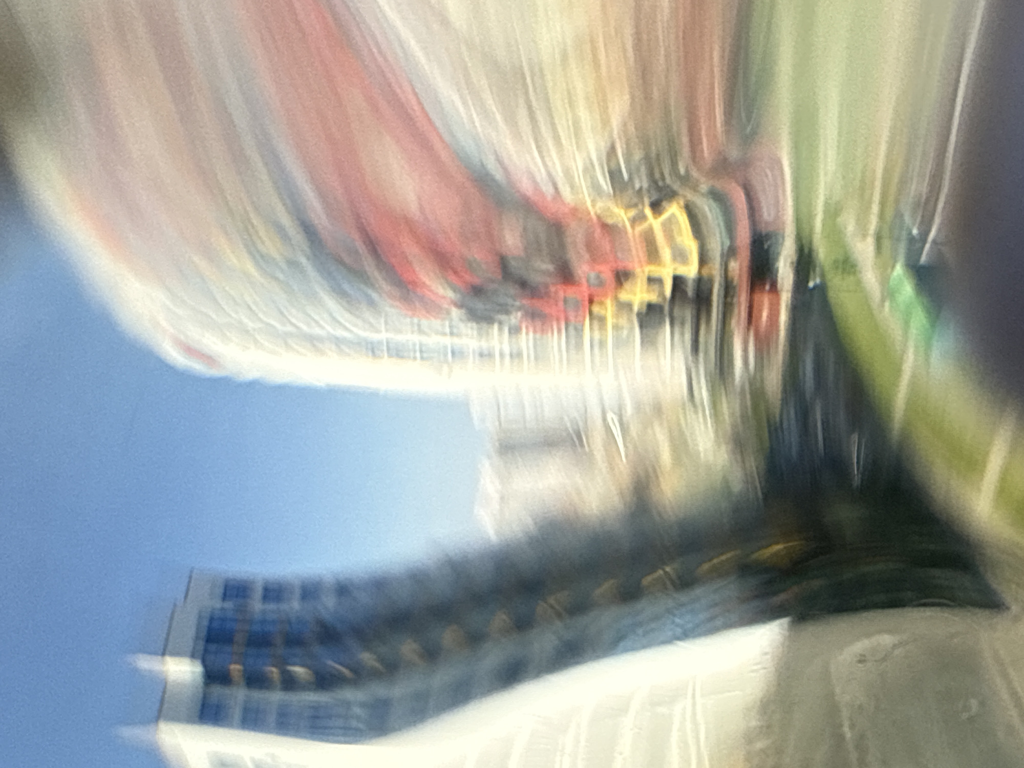 The image depicts a motion-blurred view of a cityscape, likely Minsk, Belarus. The dominant elements are tall buildings, one on the left with blue-tinted windows and a white facade, and another on the right with a curved red and yellow structure. The background is a clear blue sky. The foreground shows a blurry green area, possibly a park or lawn, and dark patches that could be pavement or shadowed areas. The motion blur suggests the photo was taken from a moving vehicle, creating streaks of light and color that abstract the urban environment. No people or specific actions are discernible due to the blur.