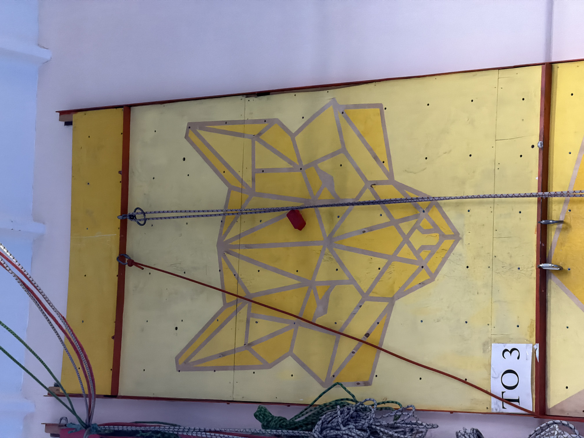A brightly lit, indoor scene shows a yellow climbing wall with a geometric bear head design. The wall is divided into sections by dark red beams. Various climbing ropes and chains hang across the wall. A white sign labeled "TO 3" is affixed to the lower right corner. The wall is situated indoors. While the location in Berezniki, Russia is noted, no specific elements in the image confirm this geolocation.
