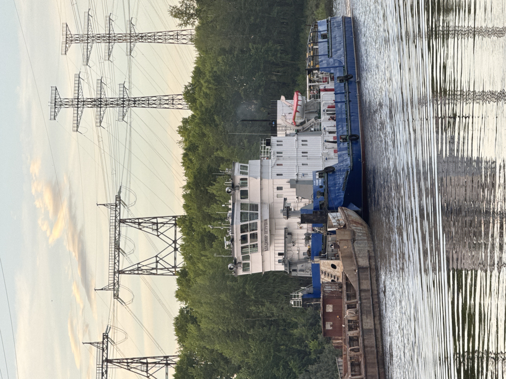 Two barges are being pushed by a tugboat down a river. The tugboat has a white superstructure with the name "ВЛАДИМИР ПЕТРУШОВ" printed in Cyrillic on its side. The barges are rusty and appear to be carrying cargo, although it is not clearly visible. In the background, there are tall power pylons and a dense line of green trees. The water is calm and reflects the sky and the power lines. The scene appears to be set in the city of Chaikovsky, Russia, given the presence of the power pylons which are characteristic of the area near the Votkinsk Hydroelectric Power Station.