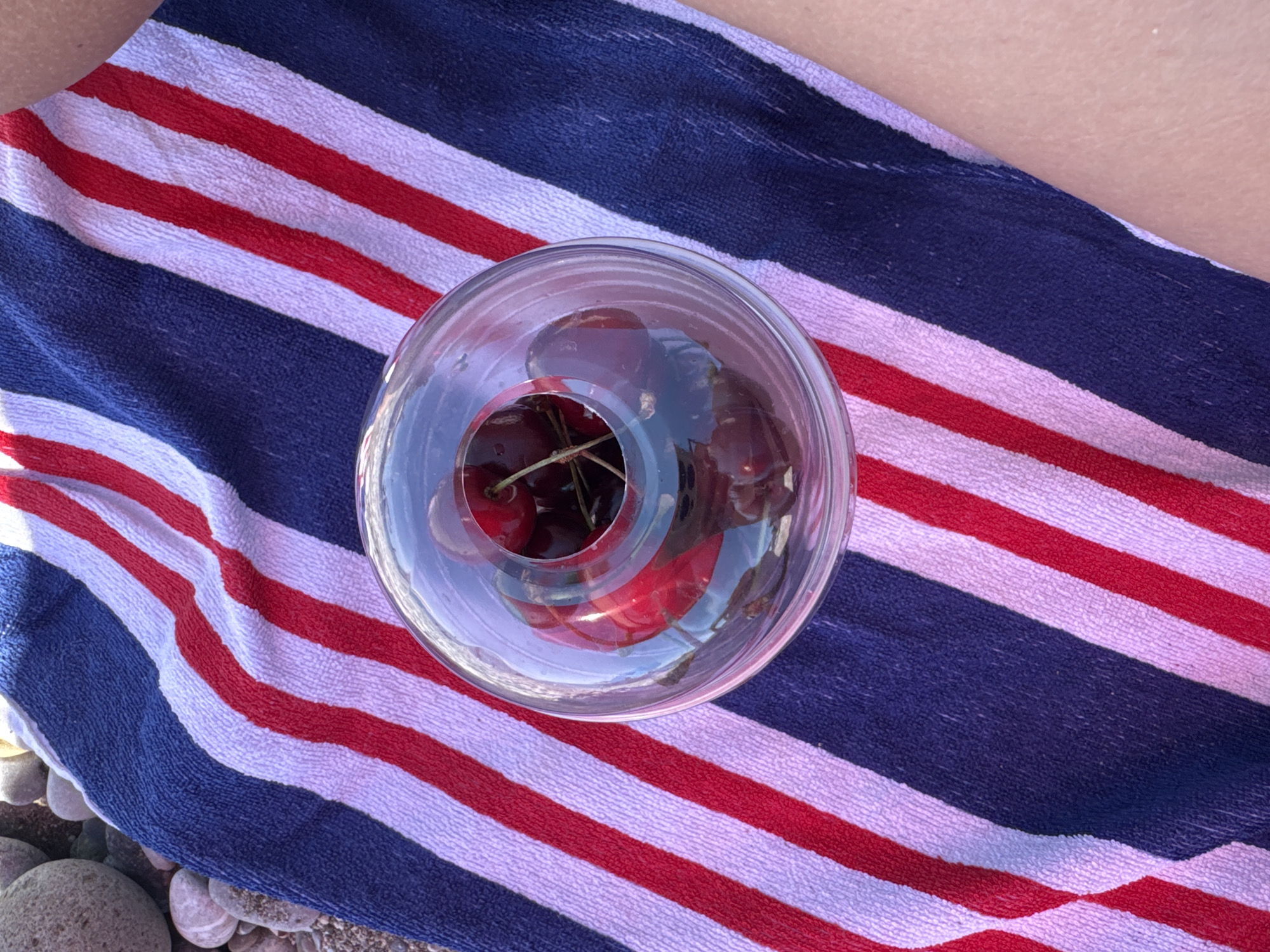 A close-up, top-down shot shows a glass bowl filled with dark red cherries sitting on a striped towel. The towel has thick, diagonal stripes of red, white, and navy blue. To the left of the towel, a scattering of smooth, grey beach pebbles is visible, suggesting a beachside location. To the right, a portion of a person's bare arm, with visible skin texture, rests on the towel. The cherries are deep red, some with stems still attached, and appear plump and ripe. The glass bowl is clear and reflects the light and the colors of the towel. The overall scene evokes a relaxed, summery atmosphere.