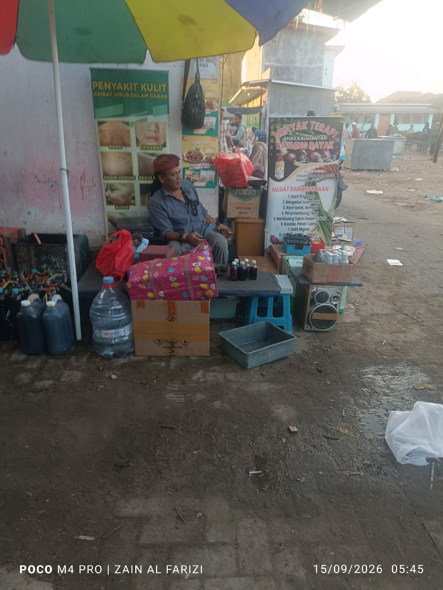 In the city of Arosbaya, Indonesia, a street vendor sits at a stall under a yellow and green umbrella. The stall displays various products, including bottles of liquid, a large water jug, and several boxes. A sign behind the vendor advertises "Minyak Terapi Khas Kalimantan Bawang Dayak," listing its benefits such as treating boils, itching, and stomach issues. Another poster on the stall's wall depicts images of skin conditions like acne and scabies. The ground around the stall is muddy and wet, suggesting recent rain or a damp environment. Several plastic containers and a speaker system are also visible. In the background, other stalls and people can be seen, indicating a market or busy public area. The overall scene depicts a typical street vending operation in Indonesia.