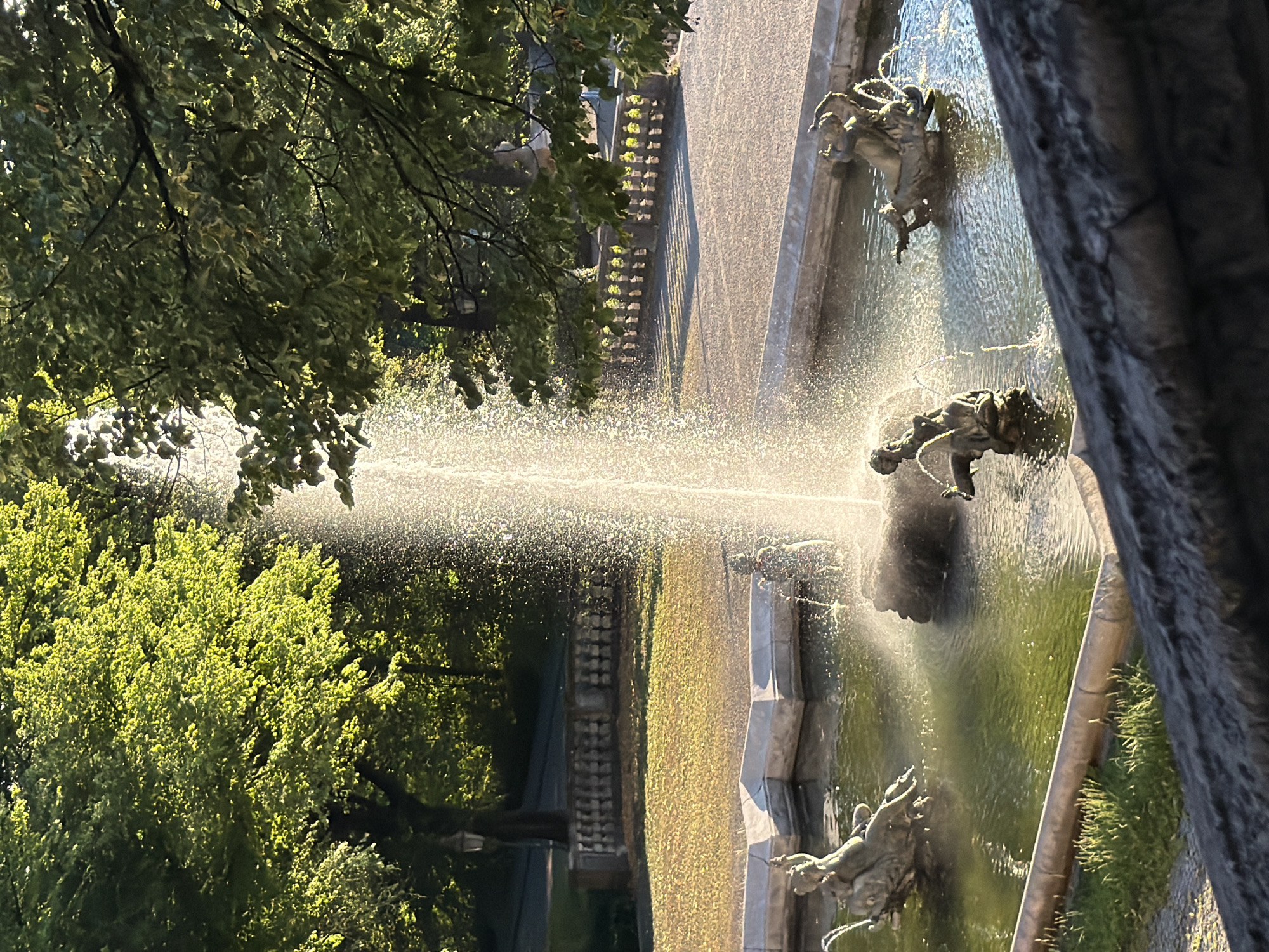 The image depicts a fountain in the Englischer Garten, Munich, Germany. The fountain features several bronze sculptures of figures, some appearing to be children or mythical beings, within a circular pool. Water sprays upward from the center of the pool, creating a misty effect that catches the sunlight. Lush green trees with dappled sunlight filtering through their leaves frame the upper portion of the scene. In the background, a stone balustrade runs along a pathway, with a lawn covered in fallen leaves visible behind it. The overall atmosphere is serene and illuminated by the warm glow of sunlight, suggesting it was taken in the late afternoon or early morning.