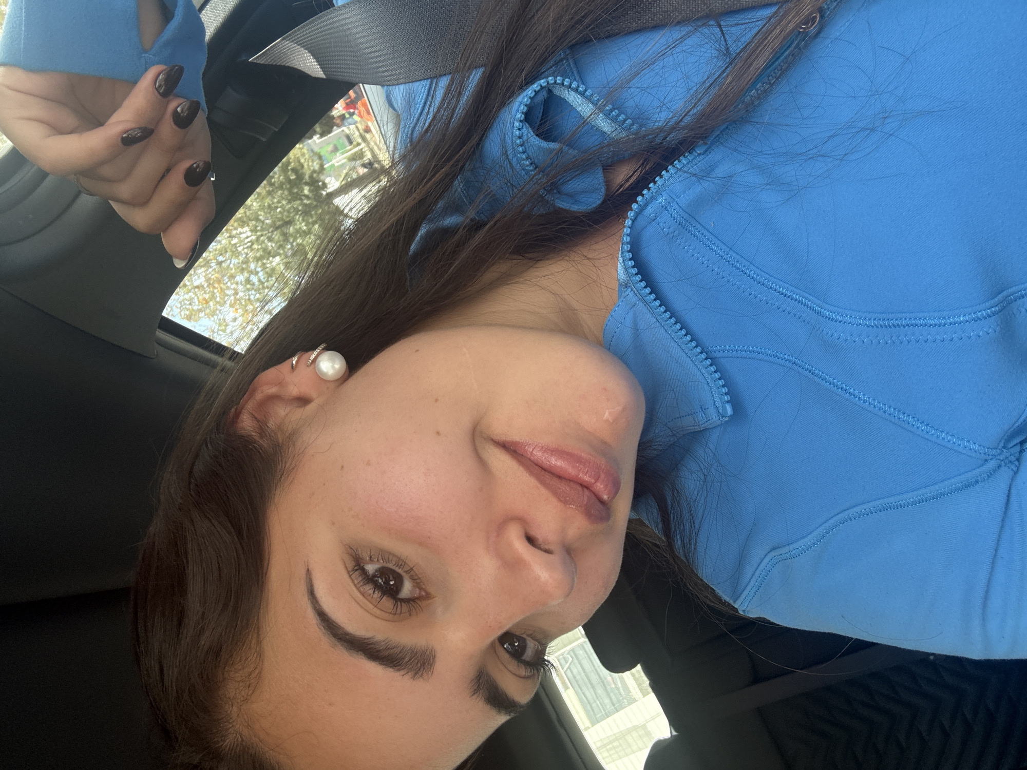 A medium close-up shot shows a woman in a blue zippered athletic jacket with her dark hair flowing around her shoulders. She has dark eyebrows and a pearl stud earring is visible in her left ear. She is wearing a seatbelt. Her left hand is visible in the upper left corner of the frame, displaying dark nail polish.
The background includes a glimpse of the outdoors through the car window, suggesting a location in Puebla, Mexico, due to the style of architecture.