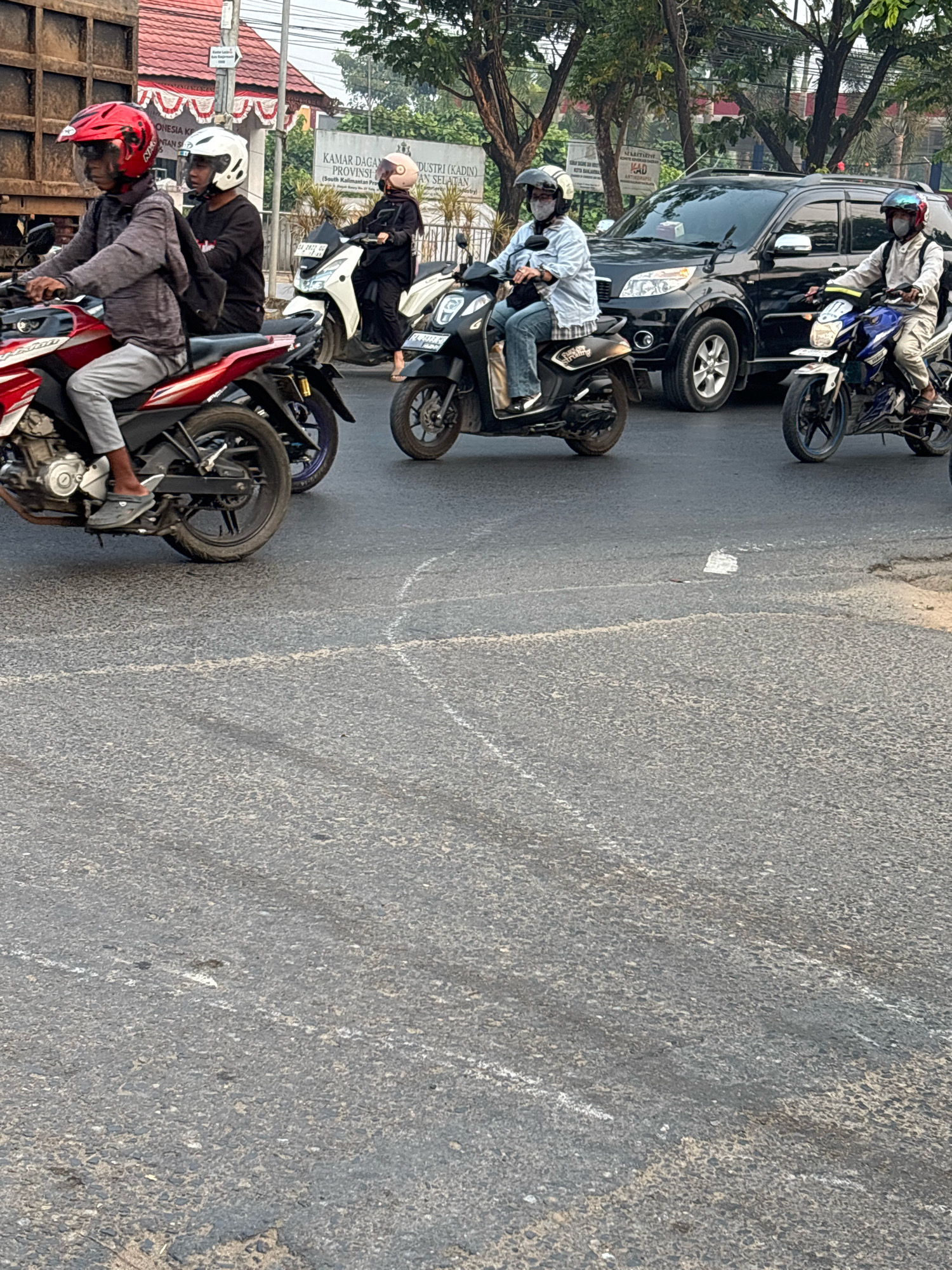 In Banjarmasin, Indonesia, at a road intersection, several individuals are seen riding motorcycles amidst traffic. The scene depicts a typical urban traffic situation with multiple motorcycles and a black SUV. The road surface is asphalt with visible white lane markings. In the background, there are buildings and trees, indicating an outdoor urban environment. One person in the foreground is wearing a red helmet and a grey jacket, riding a red and black motorcycle. Another rider in black with a white helmet is behind them. Further down the road, a person in a black outfit and a light pink helmet is on a white motorcycle. To the right, another individual in a light blue shirt and jeans, wearing a white helmet and mask, rides a black scooter. A person in a grey jacket and a blue helmet is on a blue and white motorcycle. The overall impression is of a busy street with vehicles in motion.