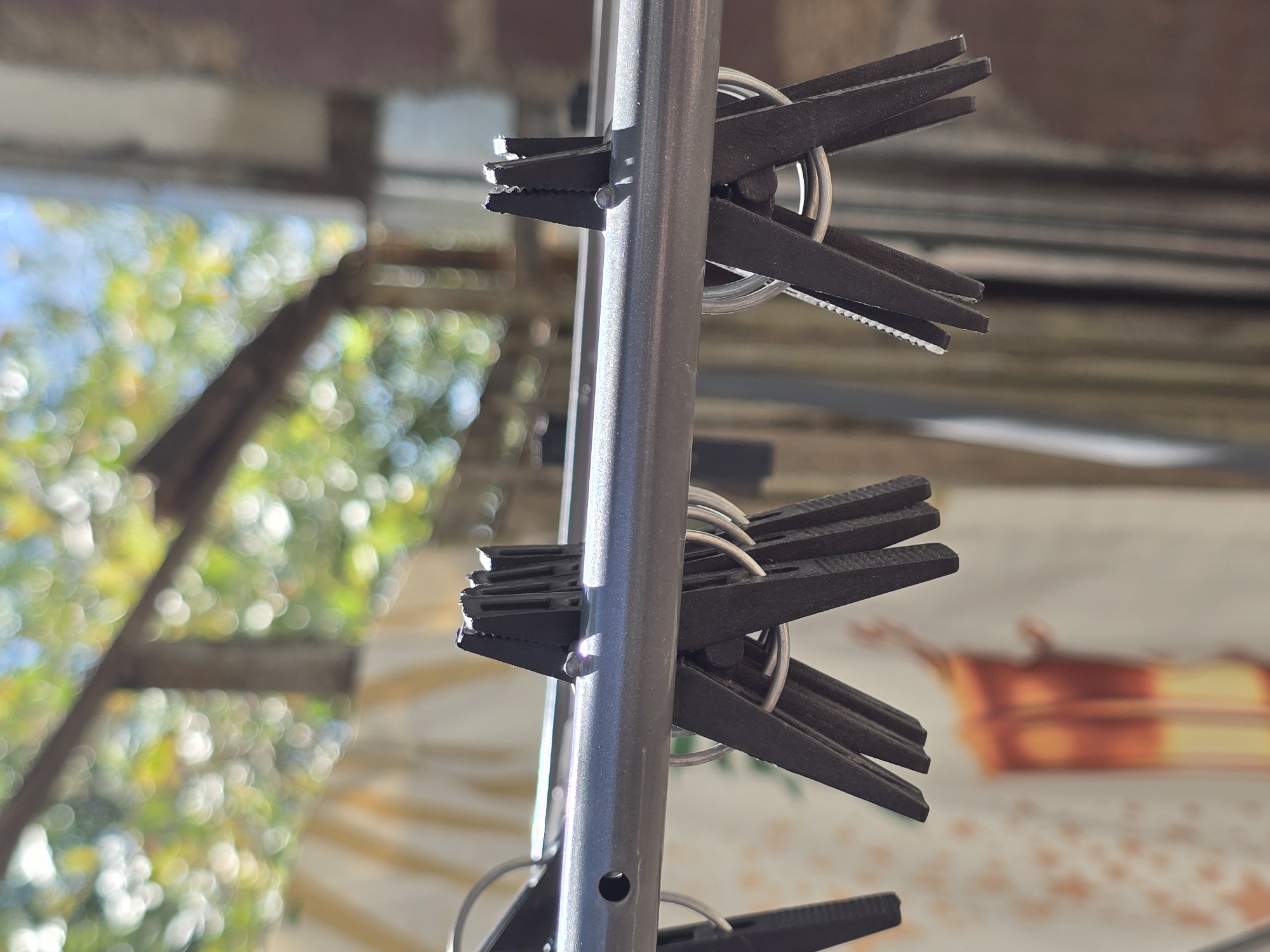 The image displays a close-up, low-angle view of black plastic clothespins hanging from a horizontal metal rod, possibly part of a clothesline. The clothespins are clustered in groups, with some showing their serrated gripping surfaces. The background is blurred, featuring sunlight filtering through green foliage and a hint of a blue sky, suggesting an outdoor setting on a sunny day. There are also out-of-focus elements that could be laundry or other objects hanging in the vicinity, and a brick wall is visible to the right. The scene lacks any discernible human presence. The context of Qaratau, Kazakhstan, cannot be inferred from the visual information provided in the image.