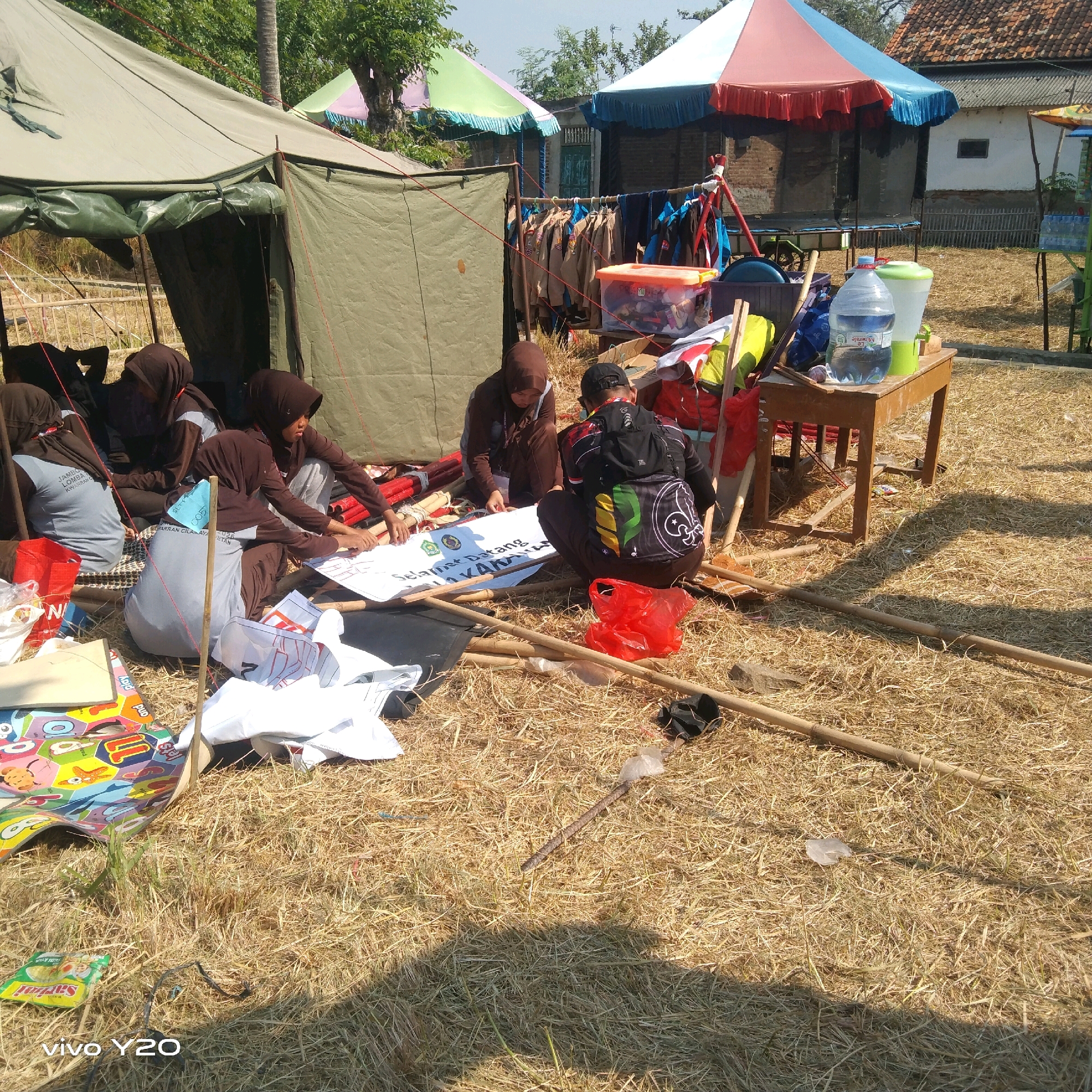 This image shows a group of young people, likely students, sitting on dry grass and working on a large white banner. They are gathered under a large, olive-green tent. The people are wearing brown hijabs and some are wearing light blue t-shirts with writing on them. The banner has Indonesian text written on it. In the background, there are colorful tents, a clothesline with uniforms hanging, and a wooden table with water bottles and other items. The environment appears to be an outdoor setting, possibly a schoolyard or a campsite, with dry straw covering the ground. The image seems to have been taken during the day, under bright sunlight.
