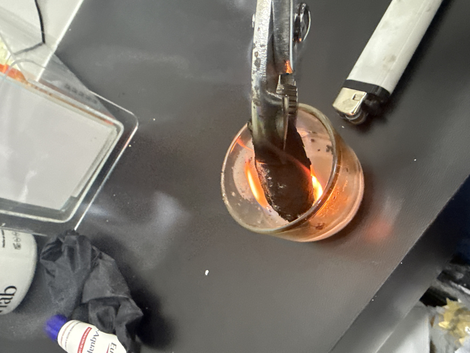 The image depicts a close-up view of a dark, possibly charred, object being held over a lit candle in a clear glass holder by a metal tool, possibly pliers or tongs. A white lighter is lying on the surface next to the candle. Nearby, there is a transparent plastic case, a white tube labeled "ab", a pair of black gloves, and a blue-capped tube labeled "Aquaphor." The objects are sitting on a gray or dark-colored surface.
It is not possible to determine the specific location of San Bernardino Tlaxcalancingo, Mexico from the image. There are no identifying elements in the image indicating the location.