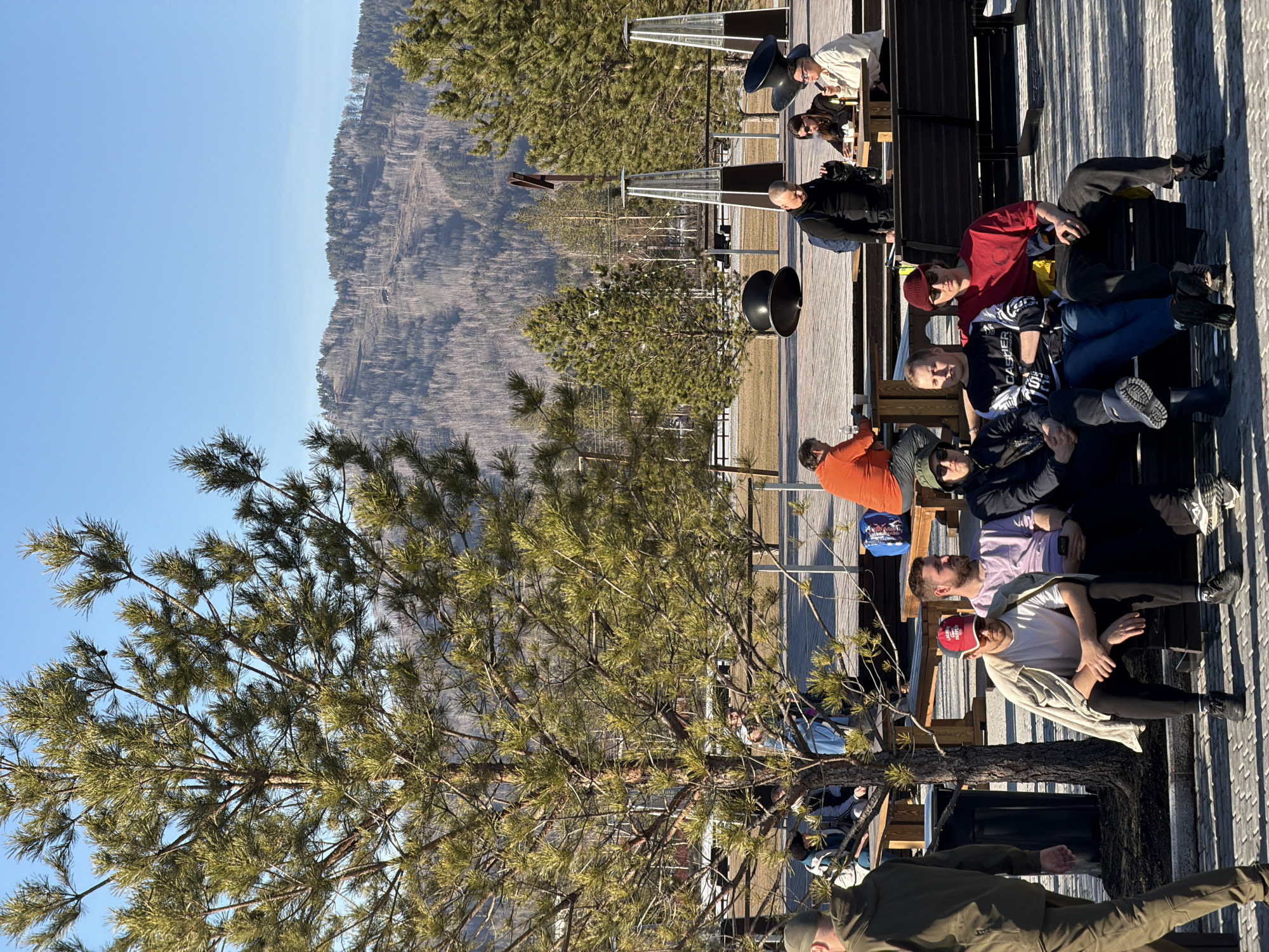 A group of approximately 14 people sit on benches on a wooden deck, overlooking a mountainous terrain under a clear blue sky. The scene is situated in Озёрное, Russia. The people are casually dressed and appear to be relaxing and engaging in conversation. Several industrial-looking speakers and heating units are visible in the background, along with pine trees.