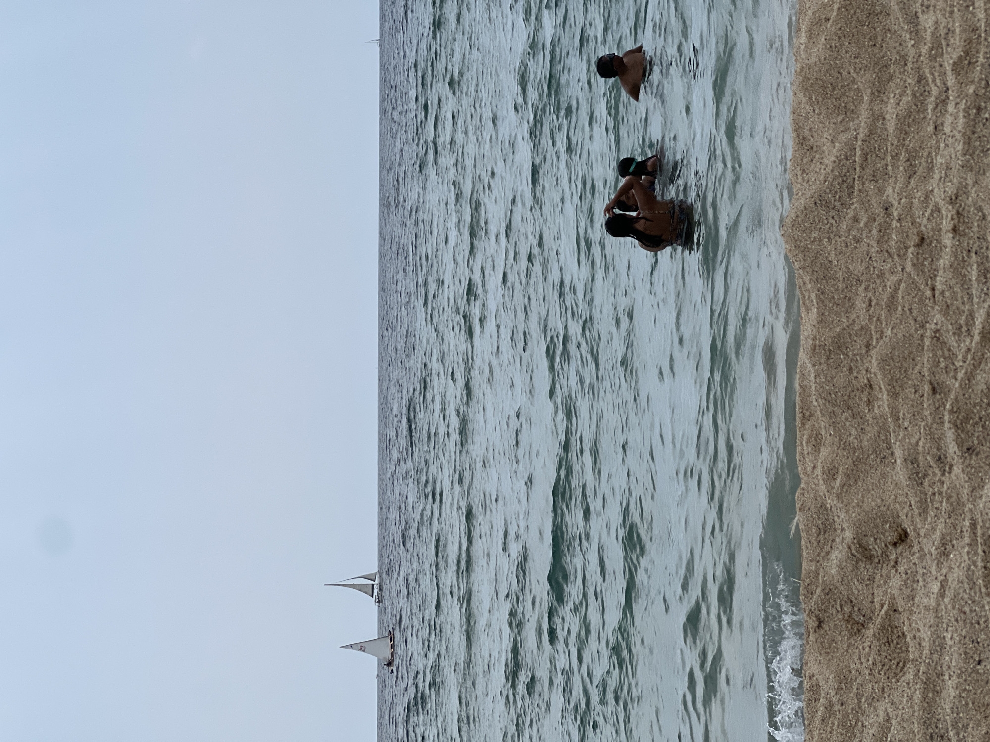 The image captures a serene beach scene in El Masnou, Spain. The foreground is dominated by a textured, light brown sand dune, with a small wave gently lapping at its base. Beyond the sand, the teal-colored sea stretches out, with moderate ripples indicating a light breeze. In the mid-ground, four individuals are partially submerged in the water. Three of them are clustered together: one person with dark hair is facing away from the camera, appearing to interact with a child, also with dark hair, who is wearing a blue swim cap. To their right, another child or young person with dark hair and goggles is visible. Further to the right, a man with short dark hair and goggles is seen from the shoulders up. In the distance, the horizon is marked by the meeting of the sea and a pale, overcast sky. Three sailboats are visible on the water. Two are closer to the left side of the frame, appearing to be part of a sailing race with one displaying a sail number "1846". A third, smaller sailboat is positioned further to the right on the horizon. The overall atmosphere is peaceful and recreational.