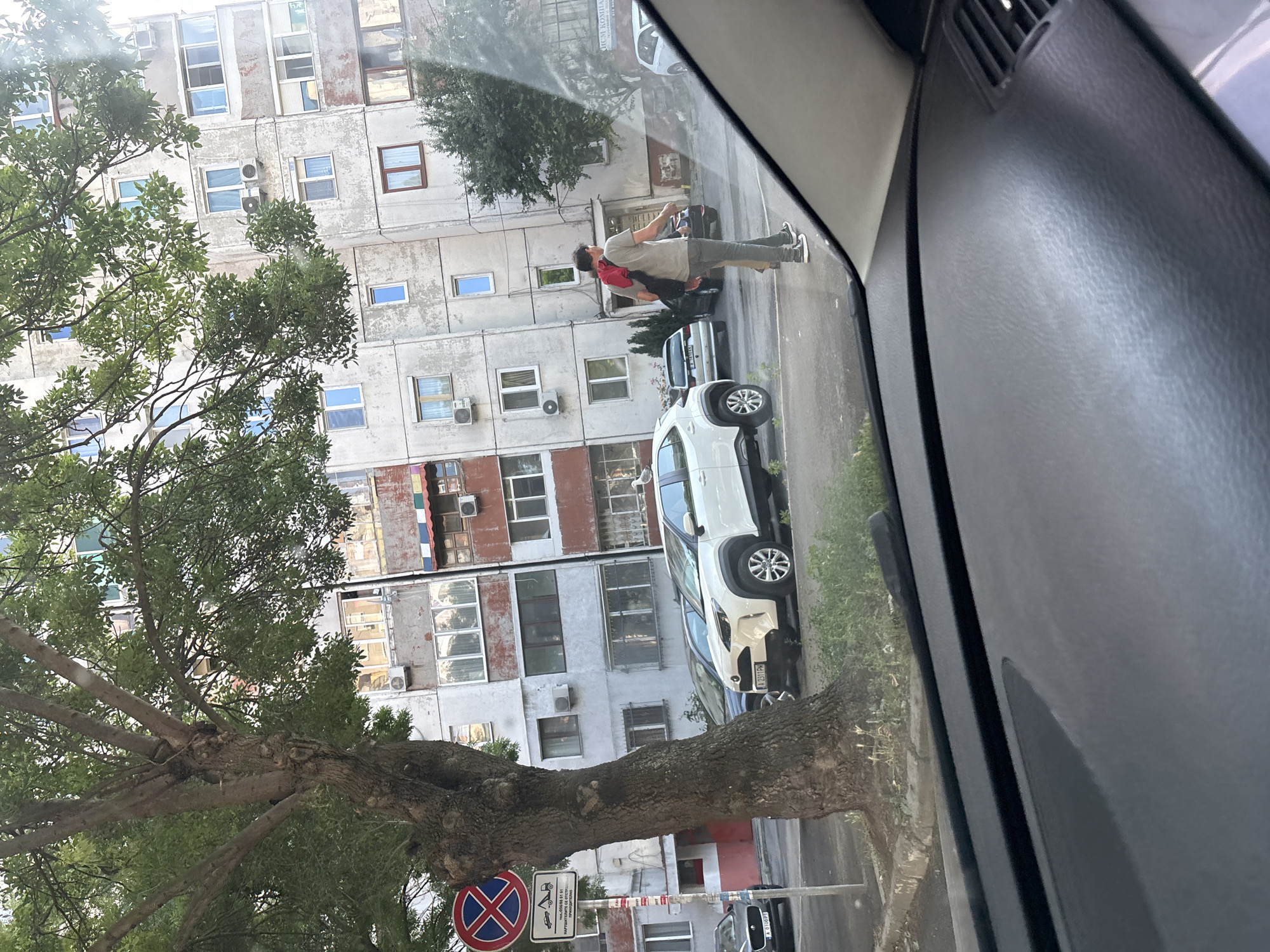 The image shows a street scene in Burgas, Bulgaria. A large tree with dense green foliage stands in the foreground, partially obscuring a tall, weathered apartment building. The building has multiple floors with numerous windows, some with air conditioning units attached. Several cars are parked along the street, including a white SUV and a dark sedan. A man wearing a red backpack and a light-colored t-shirt and pants is walking across the street. A traffic sign with a red circle and a blue cross, indicating no parking, is visible on the left side of the image. The scene appears to be taken from inside a car, looking out through the windshield. The overall environment suggests an urban setting with a mix of residential buildings and street activity.