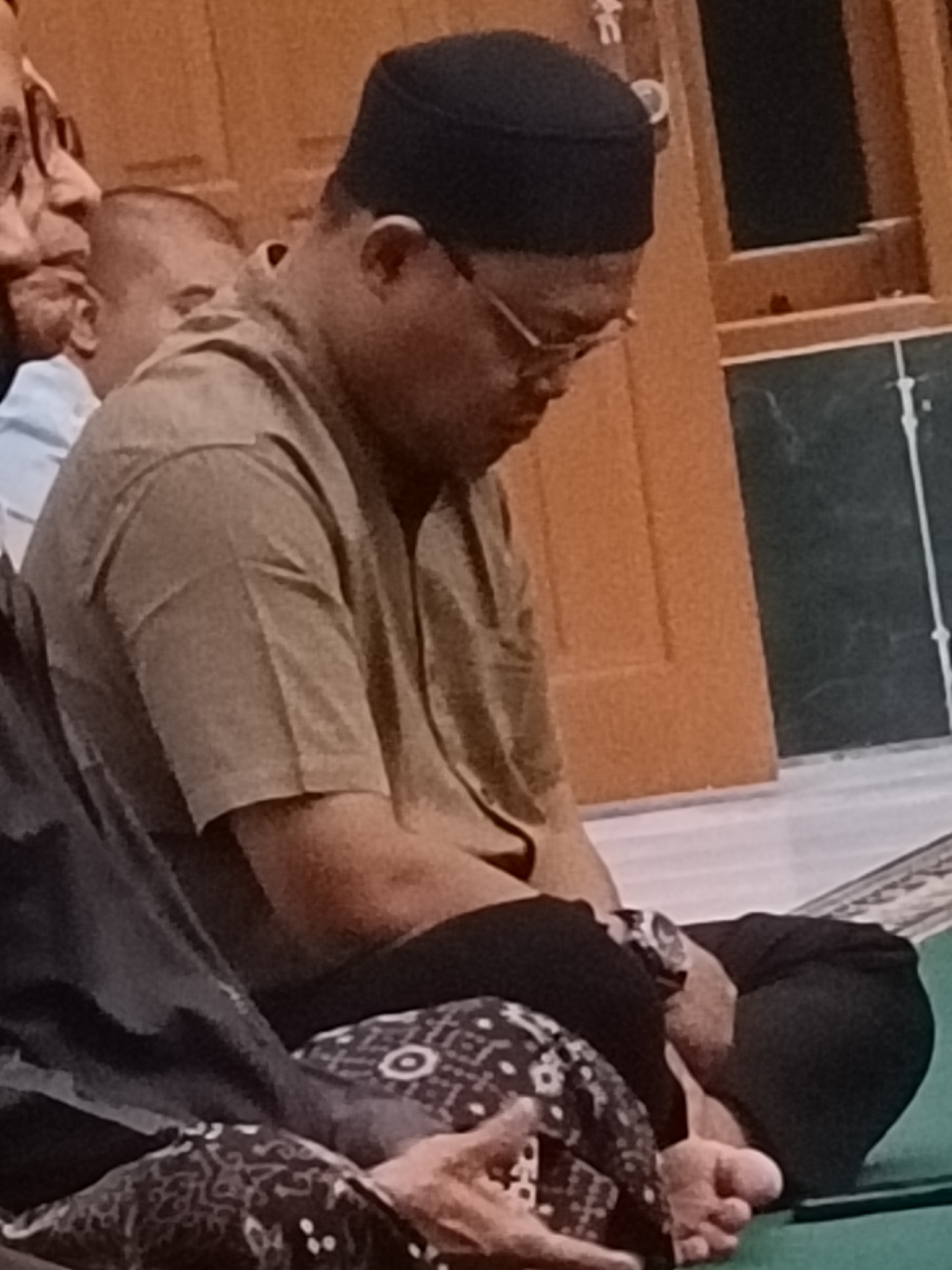 The image shows a man wearing a black cap and glasses, sitting on the floor in a room that appears to be a place of worship. He is wearing a tan collared shirt and dark pants. There are other people visible in the background, out of focus. The room has wooden paneling on the walls and a green floor covering, possibly a carpet or mat. The scene is illuminated by warm, artificial lighting. The man is looking down, with his hands in his lap. The image's angle suggests it was taken from a slightly elevated position, looking down at the seated individuals. The overall atmosphere is one of quiet contemplation or prayer. The location is identified as Kediri City, Indonesia, which suggests this is likely a mosque or a similar religious building.