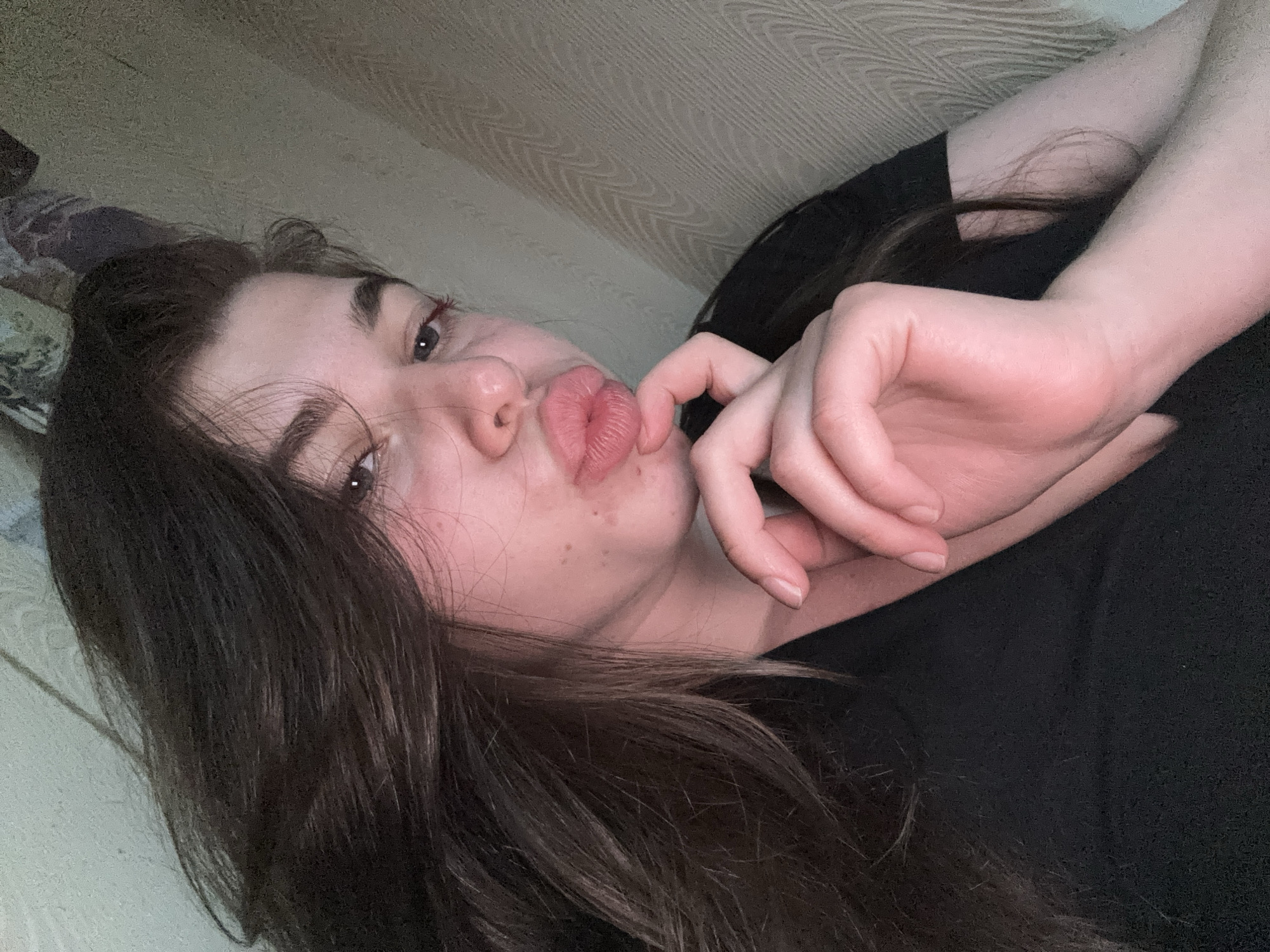 A young woman with long brown hair is lying down, facing the camera with pursed lips. She is wearing a black top. Her left hand is near her face, with her fingers lightly touching her chin. The background shows a textured surface that could be a wall or headboard. The image appears to be taken indoors with soft lighting. There is no evidence that suggests any specific location, including Мещёры, Russia.