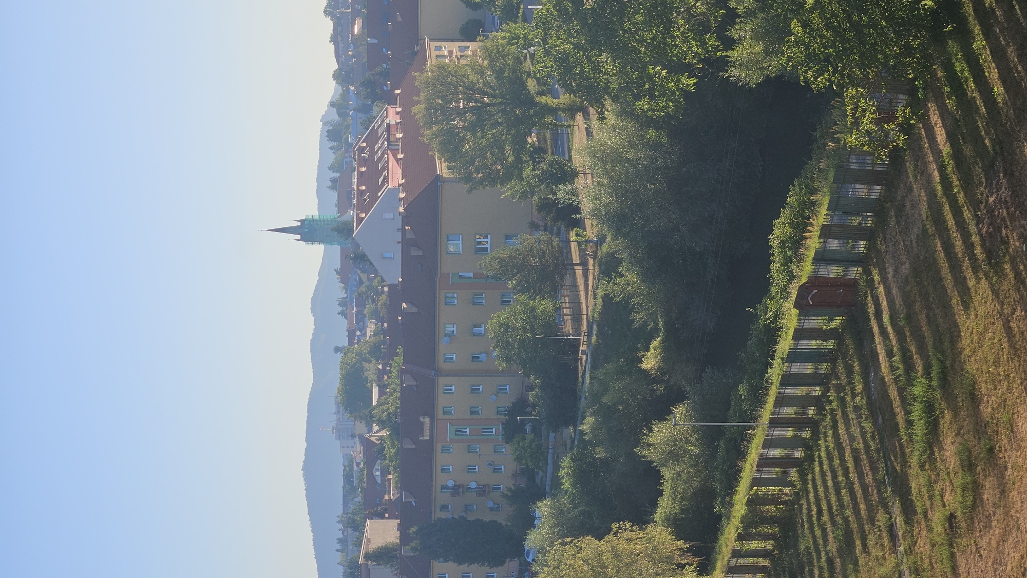 The image depicts a cityscape of Prešov, Slovakia, bathed in the warm light of early morning or late afternoon. In the foreground, a dry, grassy area is bisected by a fence constructed from vertical posts and horizontal supports, with sections painted in alternating green and brown. A small, reddish-brown gate is visible within the fence. Beyond the fence, a dense line of trees forms a natural border.
In the mid-ground, a cluster of apartment buildings with orange-tiled roofs and yellow facades dominates the scene. These buildings feature multiple windows and balconies. Scattered trees, some with lush green foliage, are interspersed between and around the residential structures. A clear, pale blue sky stretches across the background, meeting a distant range of hazy blue hills. A tall, slender building with a green, spire-like top, possibly a church tower under construction or renovation, is visible on the right side of the mid-ground, partially obscured by scaffolding. The overall impression is one of a quiet urban neighborhood embraced by nature.
