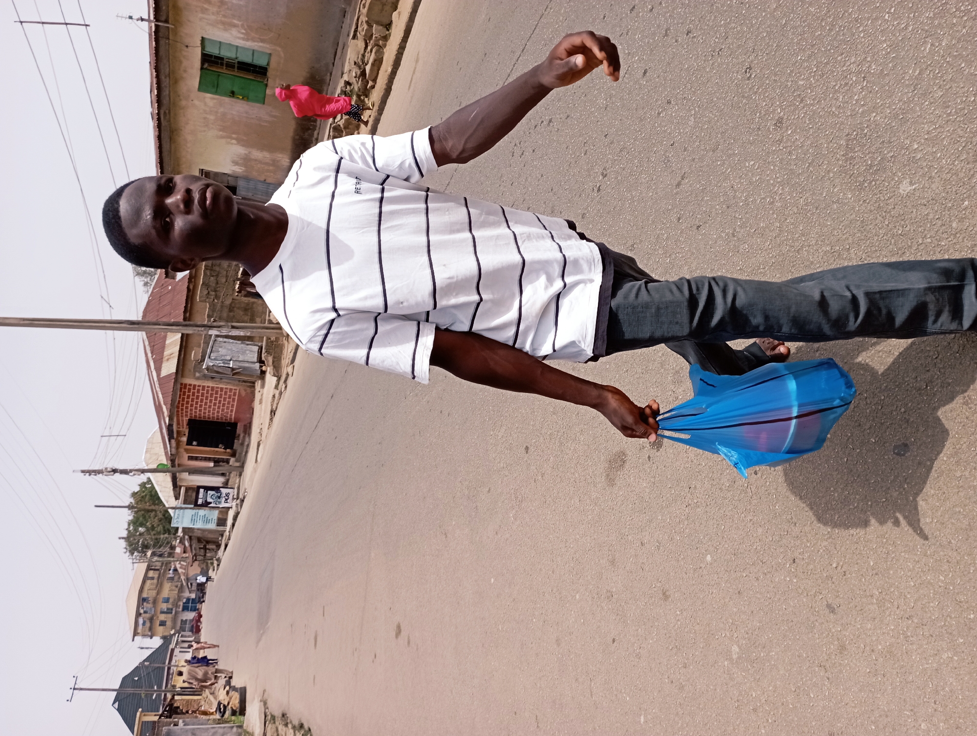 A young man stands on an asphalt road in Jos, Nigeria. He is wearing a white t-shirt with horizontal navy blue stripes, dark jeans, and sandals. He is holding a blue plastic shopping bag in his right hand and has his left arm raised. The background includes buildings, a street, a utility pole, and other people. The scene is taken during the day.