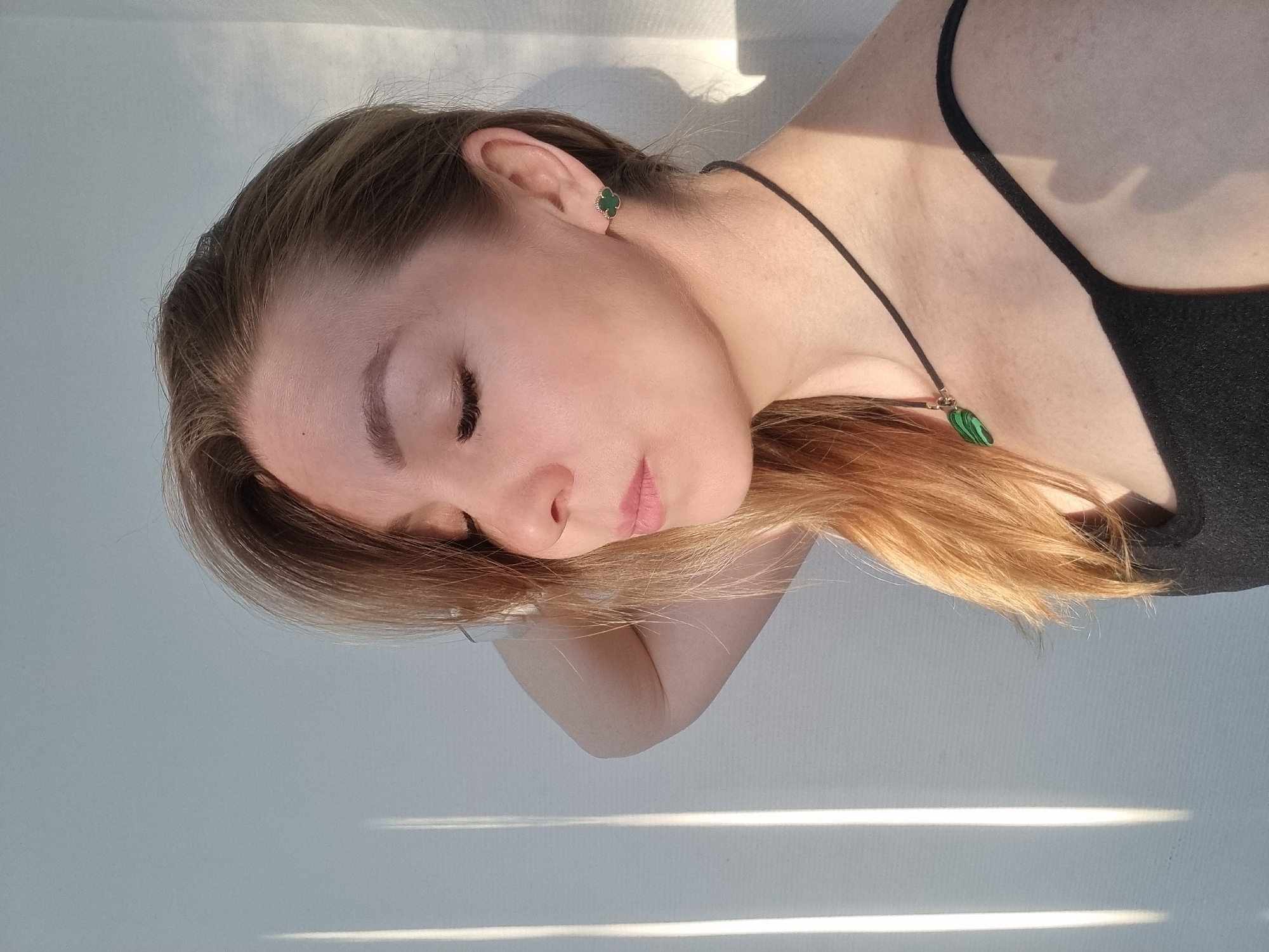 A young woman with fair skin and light brown hair is lying down, facing slightly away from the camera. She is wearing a black tank top, a black cord necklace with a green pendant, and green clover-shaped earrings. Her eyes are closed, and her face is relaxed. Sunlight streams in through what appears to be blinds, casting bright lines of light across the white surface she is resting on. The location is not evident from the image.