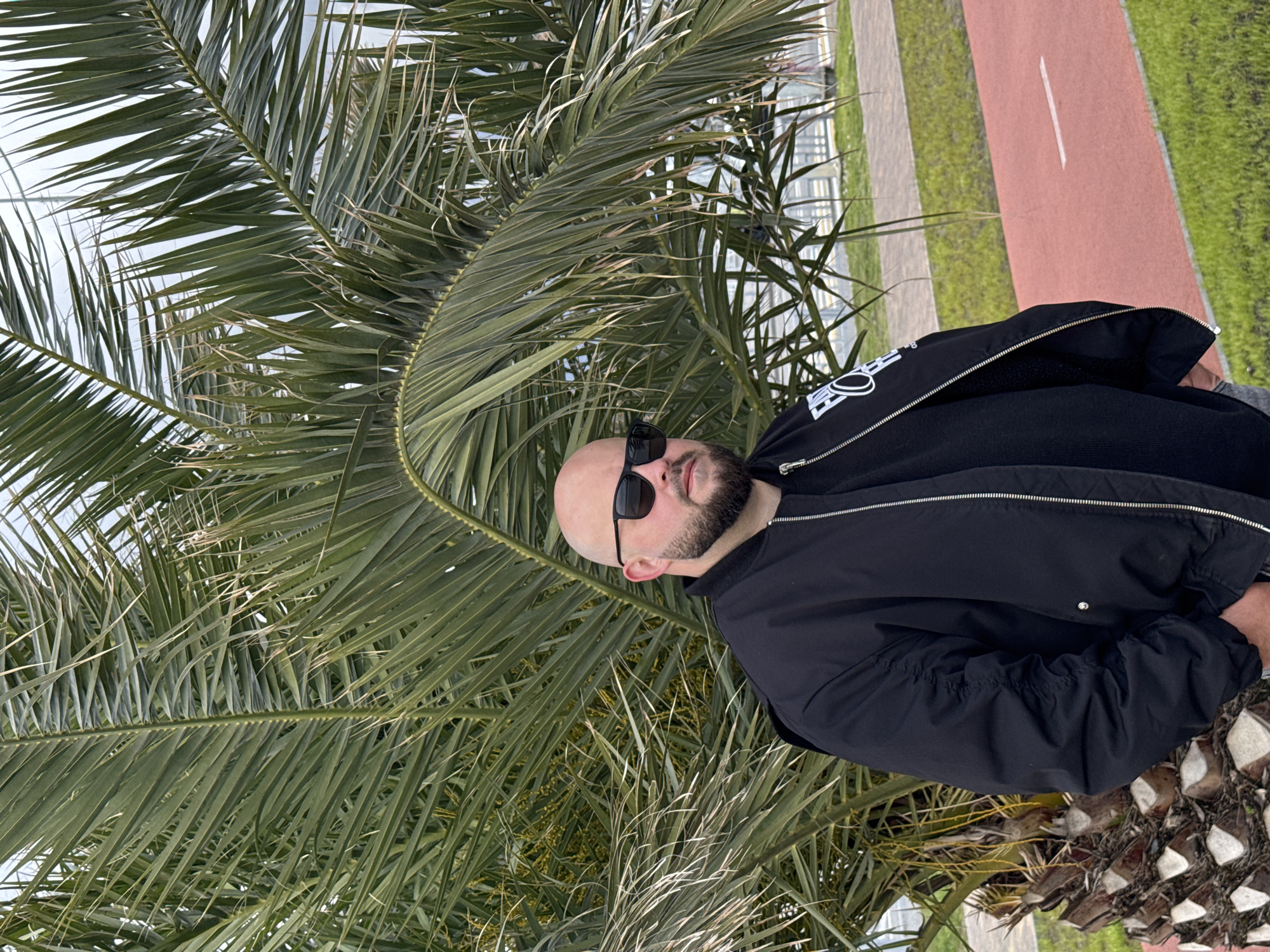 A bald man with a beard and sunglasses stands in front of a palm tree. He wears a black jacket over a black sweater. The background includes a red path, green grass, and some indistinct buildings, suggesting an urban park-like setting. The location is in the city of Sirius, Russia, though the specifics of the location within the city are not discernible.