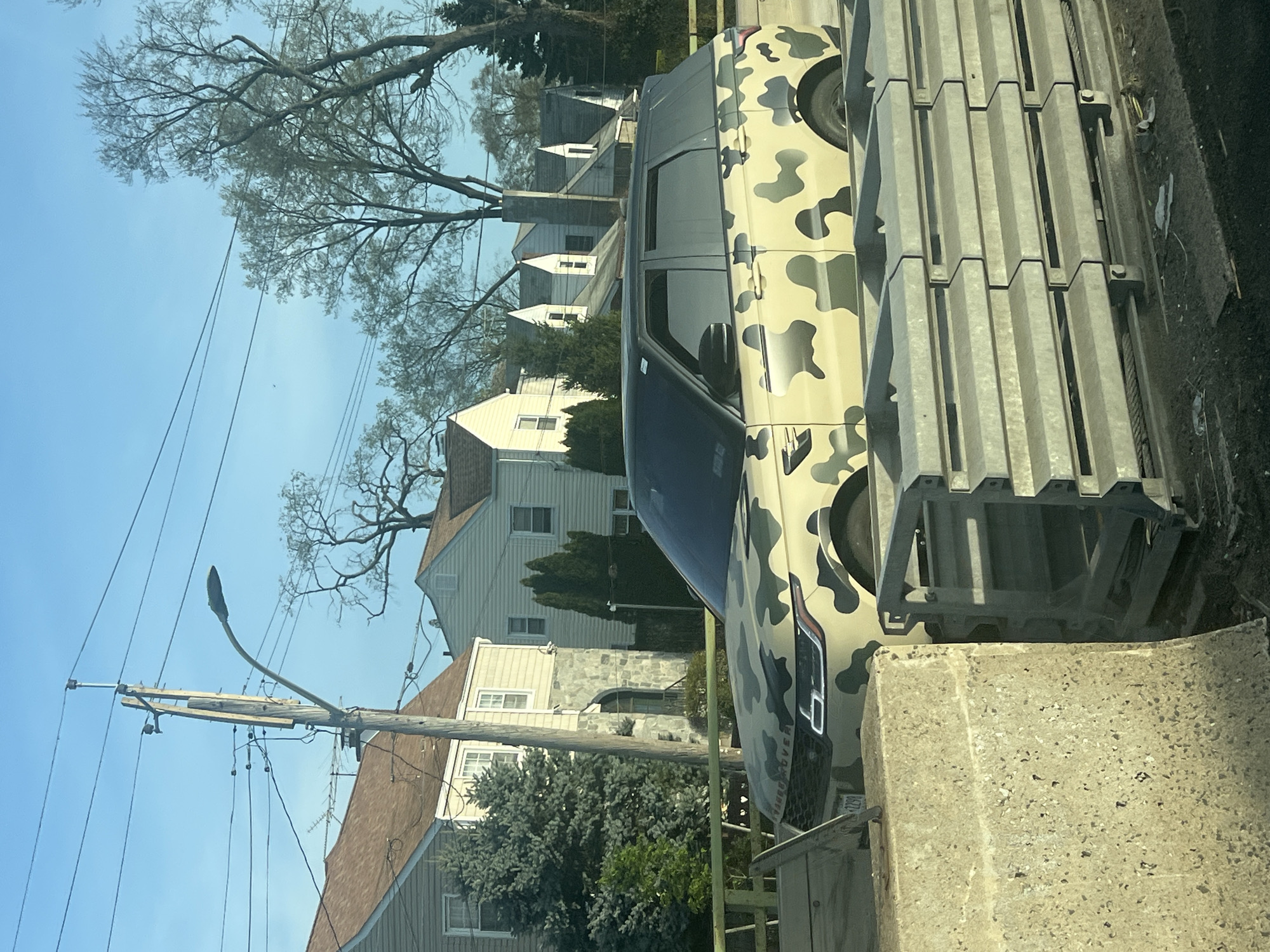 The image shows a vehicle parked on a street in a residential area. The vehicle is a Range Rover Discovery with a camouflage paint job. The license plate reads "0290". The vehicle is parked next to a concrete barrier and a metal structure. In the background are houses, trees, and a utility pole. The sky is blue. The location is the City of Yonkers, United States of America.