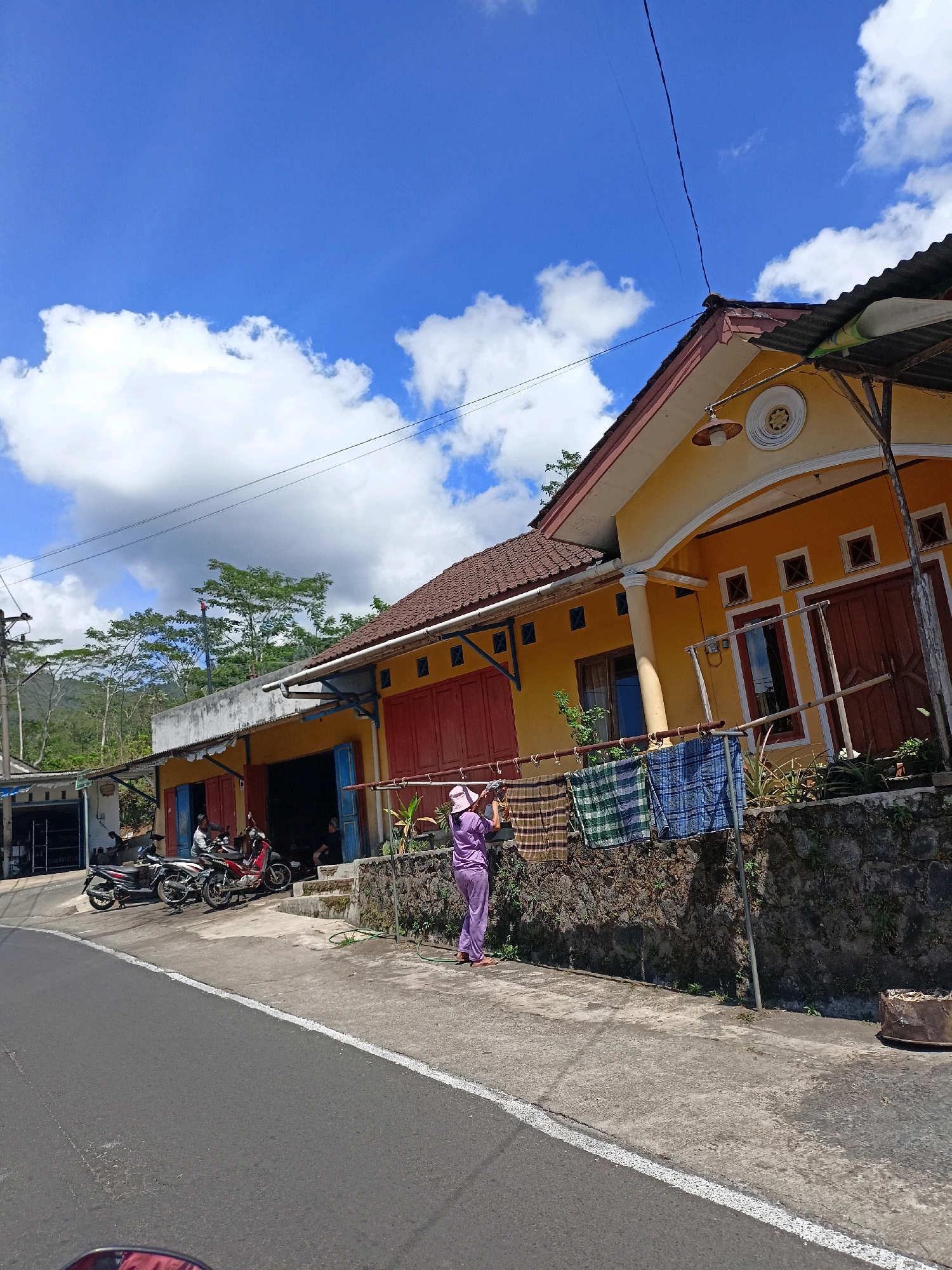The image depicts a street scene in Windusari, Indonesia, characterized by a bright blue sky with scattered white clouds. In the foreground, a paved road with a white line marking the edge is visible. To the right of the road, a stone wall separates the street from a yellow building with red doors and windows. Clothes are hanging on a rack in front of the building, and a person in a purple outfit is attending to them. The building appears to be a shop or a residence, with motorcycles parked outside another similar structure to the left. The background shows lush green trees and hills, suggesting a semi-rural or suburban environment. The overall impression is one of a sunny day in a typical Indonesian village.