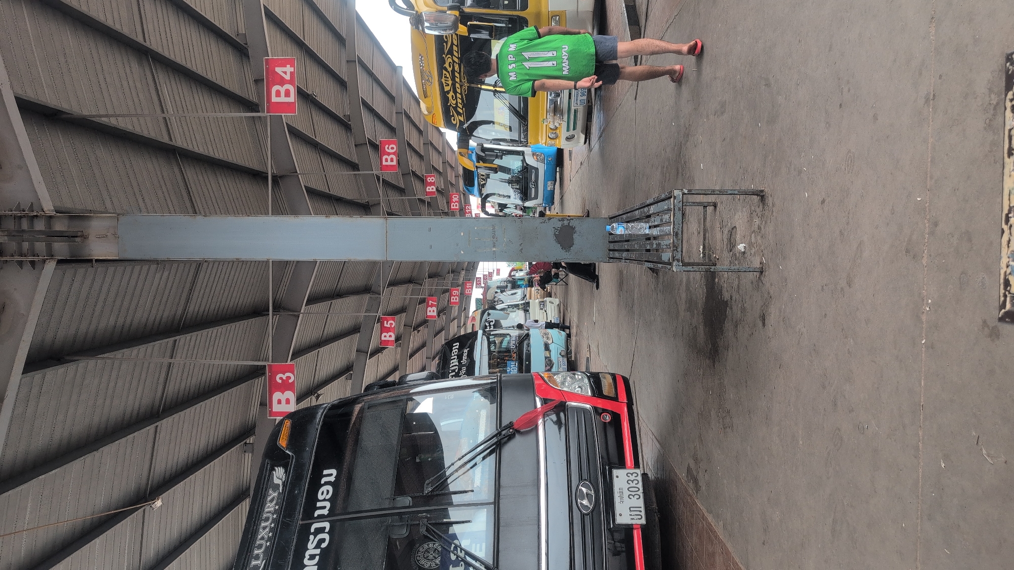 The image depicts a bus terminal in Phakhao, Laos, with several buses parked under a covered structure. The terminal appears to be outdoors, with a concrete floor and a corrugated metal roof supported by metal beams and a central pillar. Red signs with white lettering, indicating bus bay numbers such as "B3," "B4," "B5," "B6," "B7," "B9," "B10," "B11," and "B12," are suspended from the roof.
In the foreground, a black and red bus is parked on the left, displaying a license plate with the characters "UN 3033." To the right, a yellow bus with "VIP Limousine" written on it is visible. A person wearing a green shirt with the number "11" and dark shorts, along with red flip-flops, is walking away from the camera towards the yellow bus. A metal bench with a water bottle on it is positioned between the foreground bus and the yellow bus. In the background, other buses, including a light blue one, are parked, and several people are seen near the buses, some appearing to be passengers or staff. The overall environment suggests a functional transportation hub.