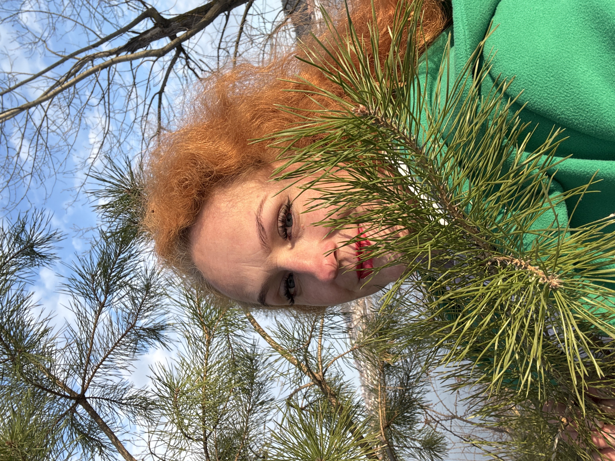 A low-angle, tilted shot depicts a woman with red hair, positioned behind pine branches. She wears a green garment, potentially a coat or jacket. Her face is visible between the pine needles, and she is looking downward. The background shows bare tree branches and a blue sky with light clouds. The location is identified as Noviy, Russia.