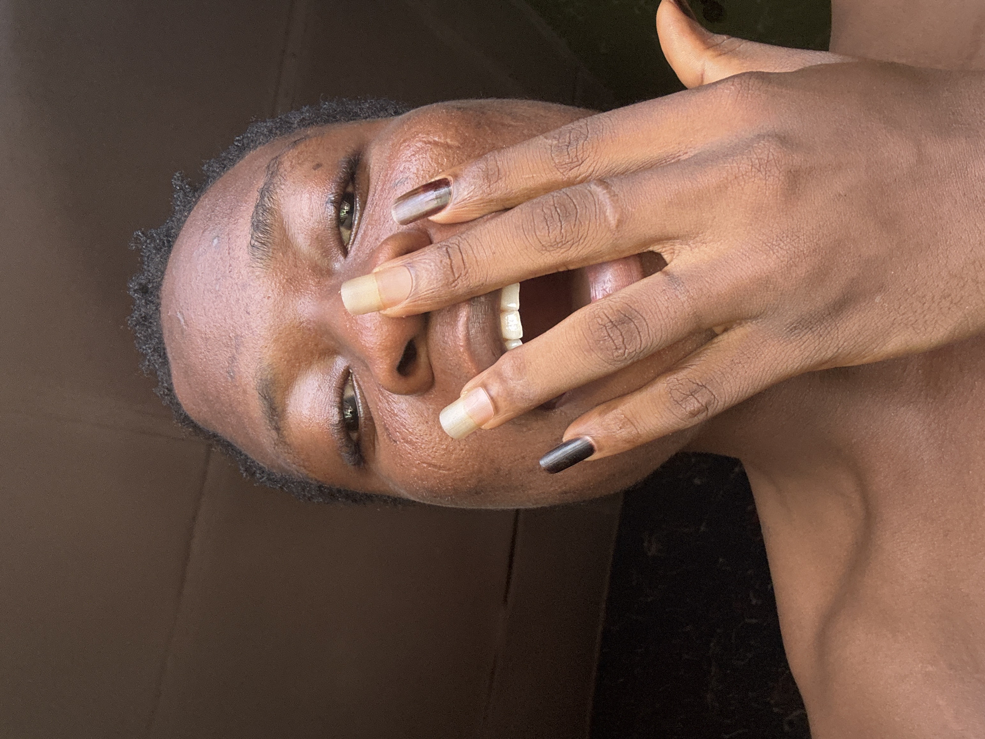 A close-up, tilted, indoor shot of a person of color's face with a hand partially covering it. The person is smiling, revealing teeth. Their eyes are open. The fingers of the hand have painted nails with two different colors. The background is ambiguous, with dark tones and hints of light. The location appears to be indoors, but without further context, the city and specific setting are unknown.