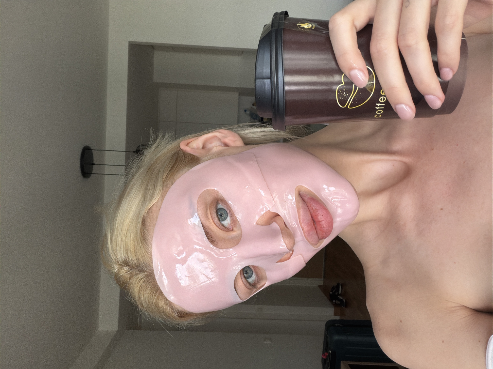 A woman with a pale complexion wears a pink sheet mask and holds a brown coffee cup labeled "coffee." She has light blue eyes and blonde hair visible around the mask. The scene is indoors, with a white wall and a dark-colored object resembling a wall-mounted rack visible in the background, along with glimpses of an open doorway. A suitcase is partially visible in the lower right corner. The location is indeterminate and cannot be ascertained as Belgrade, Serbia, from the visual information.