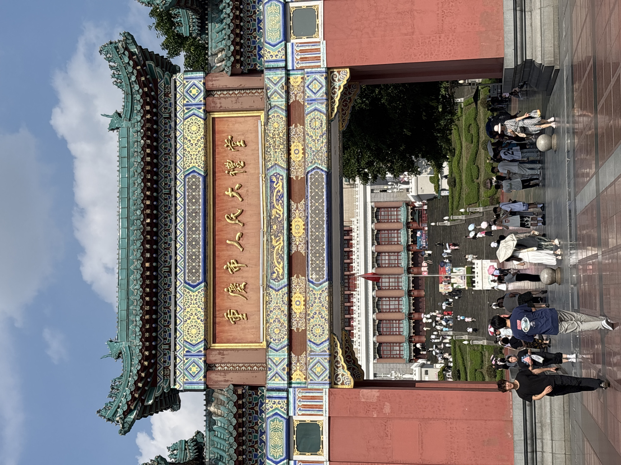 The image shows the entrance arch to the Chongqing People's Great Hall in Yuzhong District, China. The arch is ornate, with red pillars and a green, tiled roof adorned with dragon figures. Golden Chinese characters are inscribed on a plaque above the entrance. Through the archway, a wide set of stairs leads up to the grand building, which has a classical facade with numerous windows and columns. A red flag flies on a flagpole to the right of the stairs. A crowd of people, many holding umbrellas, are ascending the stairs or gathered at the base. In the foreground, several individuals are walking on a paved plaza. The sky above is blue with scattered white clouds.