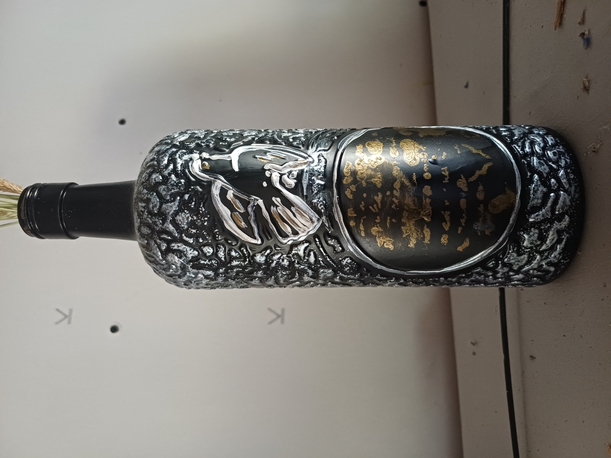A decorated black glass bottle, likely a wine bottle, stands upright on a light-colored surface. The bottle is adorned with a textured, silvery-white relief pattern resembling tree bark. In the upper half of the bottle, a white and gold depiction of a bird is prominently displayed. Below the bird, a circular emblem features a black background with scattered gold accents, possibly representing text or abstract patterns. The neck of the bottle is black and narrow, topped with a black cap, and a few dry stalks of wheat or similar vegetation protrude from the opening. The background is a plain, light-colored wall with some faint markings. A thin black wire runs horizontally across the lower part of the scene. The image is shot from a slightly low angle, looking up at the bottle.