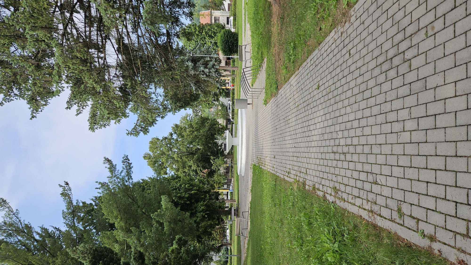 A paved pathway leads through a park in Prešov, Slovakia. The pathway, made of grey rectangular pavers, is bordered by lush green grass on the left and a line of metal benches and a bike rack on the right. In the center of the park, a large, white, multi-tiered fountain is visible. Tall, mature trees with dense green foliage surround the area, casting dappled shadows. In the background, a few buildings and cars suggest the park is situated within a residential or urban area. The sky is a pale blue with scattered clouds.