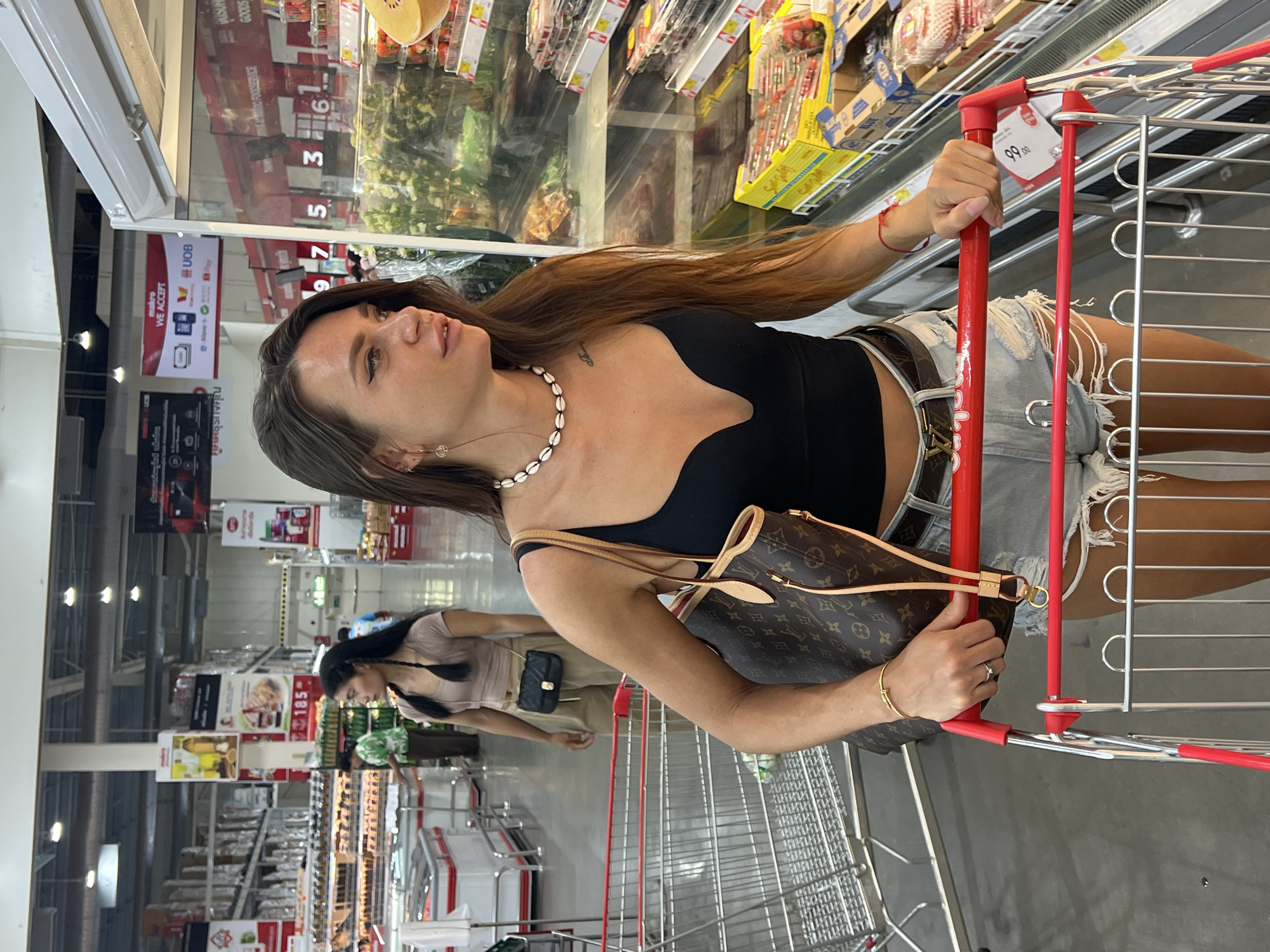 In Baan Hua Thanon, Thailand, a woman with long brown hair, wearing a black crop top, distressed denim shorts, and a shell necklace, pushes a red-handled shopping cart. She is holding the cart with her right hand, which displays a thin gold bracelet and a ring on her index finger. Beside her, a brown Louis Vuitton-patterned tote bag hangs from her shoulder. Another woman with dark hair pulled into two braids, wearing a light brown t-shirt and beige pants, is visible in the background, looking to her left. The environment appears to be a well-lit supermarket aisle with rows of shelves stacked with products, including a refrigerated display case containing fruits and vegetables on the right. The floor is a polished gray concrete.