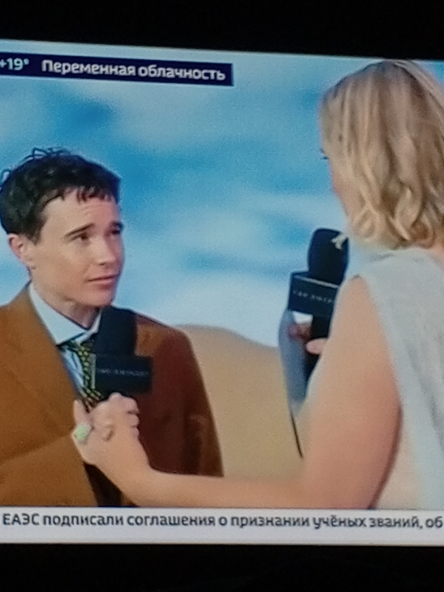 This is a screenshot of a television screen showing a news report. In the foreground, two individuals are visible. On the left, a person with short, dark hair and wearing a tan jacket over a blue shirt and a patterned tie is holding a microphone. The person is looking to their right, towards the other individual. On the right, a person with blonde, wavy hair, wearing a light-colored top, is holding a microphone. Their back is partially to the camera.

At the top of the screen, a banner displays weather information: "+19° Переменная облачность" which translates to "+19° Partly cloudy". Below the individuals, another banner displays text in Russian: "ЕАЭС подписали соглашения о признании учёных званий, об". This translates to "EAEU signed agreements on the recognition of academic degrees, on". The background appears to be a light blue sky or backdrop, possibly indicating an outdoor setting or a studio set.

The scene is set in the city of Yamashurma, Russia, as indicated by the provided context.