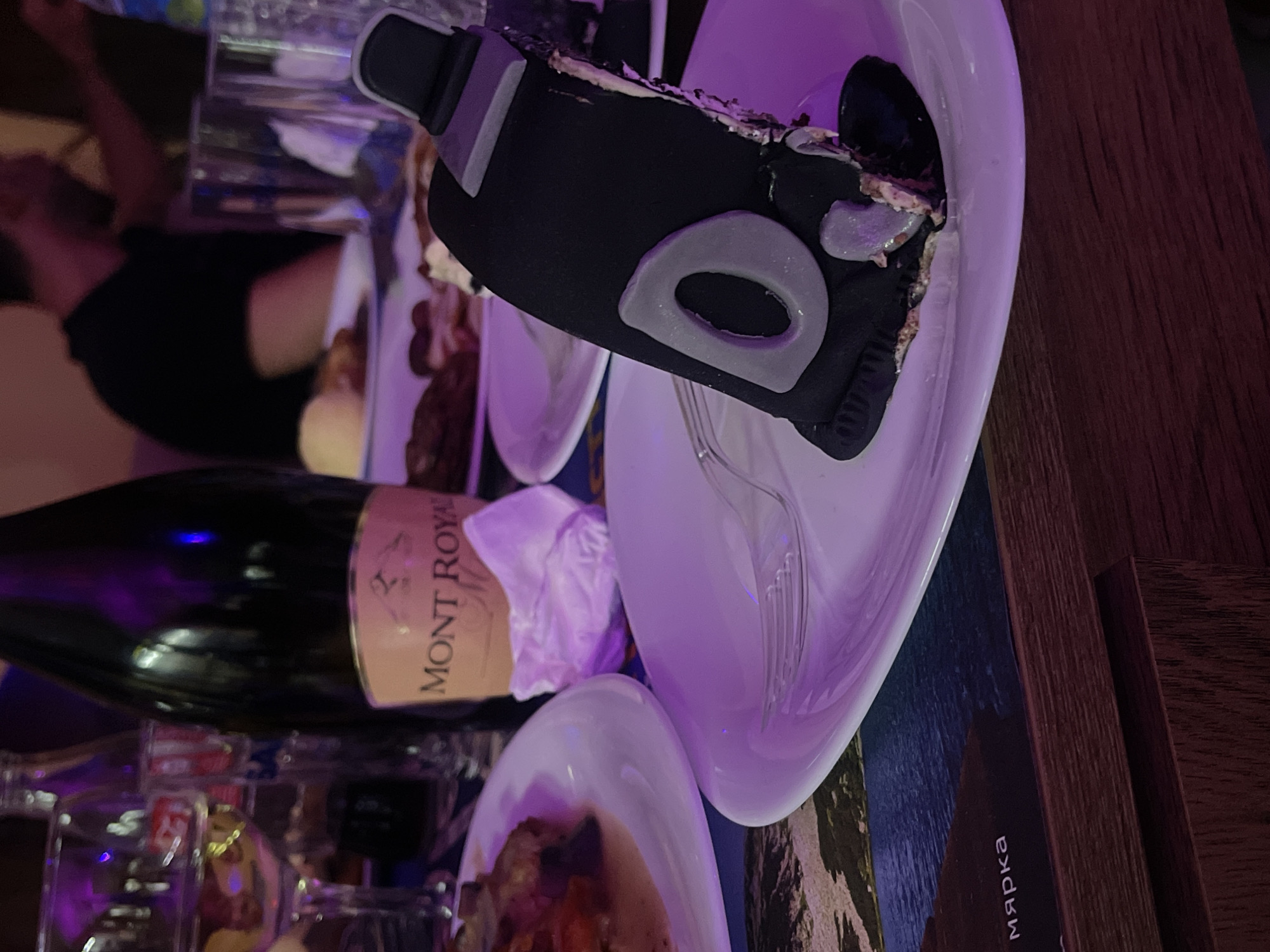 A partially eaten cake decorated as a black object resembling a VR headset sits on a white plate with a fork. A bottle of Mont Royal wine is also visible. The setting appears to be a table at a social gathering, with other plates and glasses nearby. A person's arm and part of their torso are visible out of focus in the background. The table surface has a wooden section and a section with a printed image, possibly a placemat or table covering. The city is not deducible from the image.