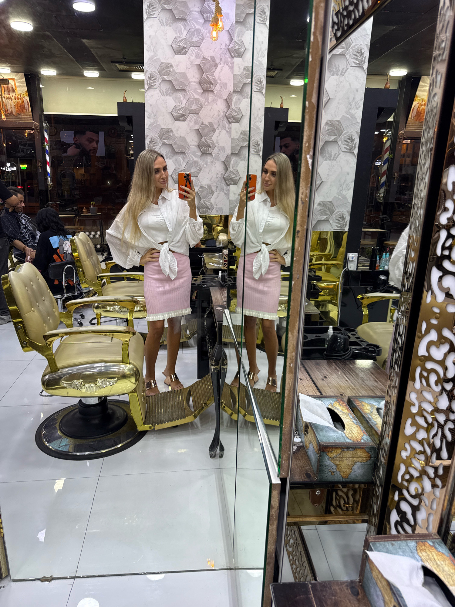 A full-body, selfie-style shot is captured in a brightly lit salon, likely in Dubai, United Arab Emirates, as evidenced by the presence of numerous opulent barber chairs. A woman with long blonde hair, wearing a white knotted shirt and a pink skirt, is positioned in the center of the frame, taking the photo with her orange-cased smartphone. Her reflection is prominently displayed in a large mirror that occupies the right side of the image.
The salon interior is characterized by its luxurious elements. Several ornate, gold-colored barber chairs with intricate detailing are arranged throughout the space. The walls are decorated with a striking hexagonal pattern in shades of gray and white, adding a modern touch. In the background, other salon patrons and staff can be observed, along with various salon equipment and decor, including barber poles and displays of hair products. The flooring is a polished white tile, reflecting the overhead lighting. To the right, a section of the mirror reveals a wooden surface with decorative tissue boxes adorned with world map designs, further suggesting a sophisticated and possibly themed establishment. The overall impression is one of a high-end grooming establishment.