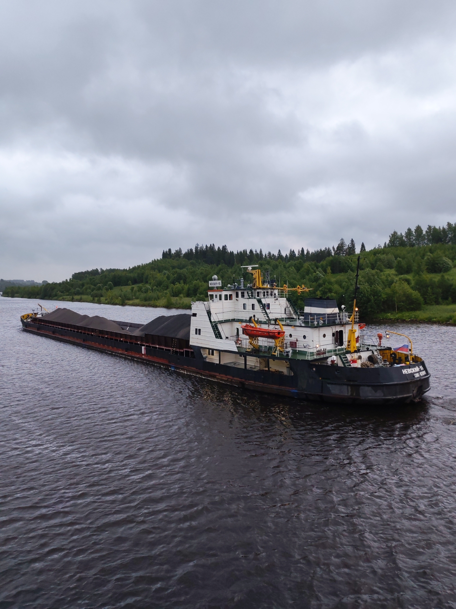 An aerial, full-shot captures a barge carrying a large quantity of dark, granular material, likely coal or similar cargo, moving on a wide, dark river. The barge is long and low, with a white superstructure at the stern. A smaller vessel, a tugboat, is attached to the stern of the barge and is pushing it. The tugboat is white with yellow accents and features various nautical equipment. A Russian flag is visible on the tugboat. The river is calm with gentle ripples. The background consists of a lush, green forest that covers rolling hills. The sky is overcast with heavy, grey clouds, suggesting a cloudy or possibly rainy day. The scene is likely in or near Alekseevskoe, Russia, given the context provided. There are no people visible in the image.