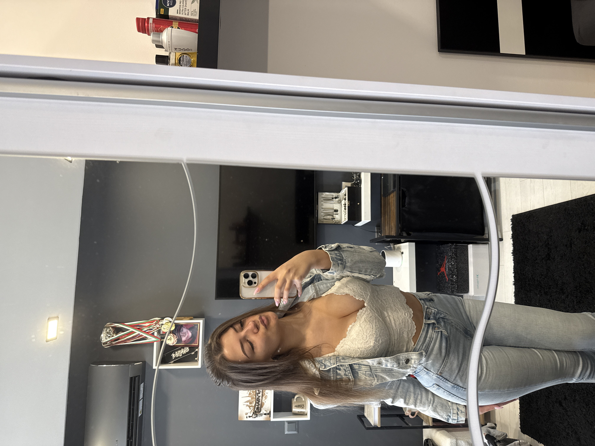 A young woman with long brown hair is taking a selfie in a mirror. She is wearing a white lace top, a denim jacket, and light blue jeans. The background includes a gray wall with a TV, shelves with various objects, a small black rug, an air conditioning unit, and a picture with medals. The location may be a modern apartment in Sofia, Bulgaria.