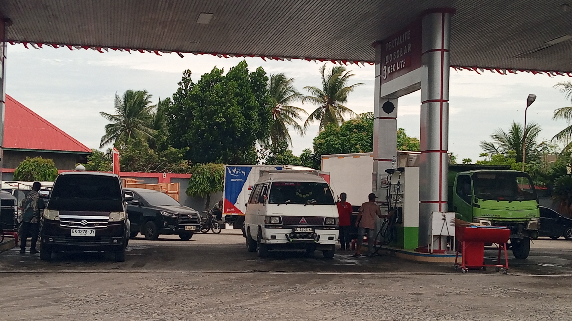 A gas station in Julikeude Dua, Indonesia, features multiple vehicles at the pumps under a covered canopy. A dark maroon van is parked at a pump on the left, with a person standing near it. To the right of this van, a dark SUV is also parked. In the center of the frame, a white van is positioned at a pump, with two individuals attending to it. Further to the right, a green truck is at a pump, with a red cleaning cart nearby. Behind the vehicles, a red-roofed building and lush green trees are visible, suggesting a tropical or sub-tropical environment. The sky is overcast. The canopy of the gas station is decorated with red and white pennant flags. Signs above the pumps indicate fuel types such as "PERTALITE," "BIO SOLAR," and "DEX LITE."