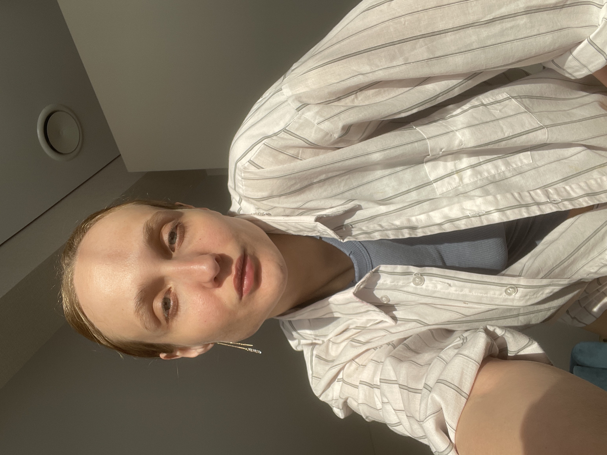 A woman is posing for a selfie indoors. She is wearing a striped white and grey button-down shirt over a blue top. The lighting is bright, casting shadows across the scene. In the background, there is a vent on the ceiling. Based on the available information, it is not possible to determine the exact location within Sepych, Russia.