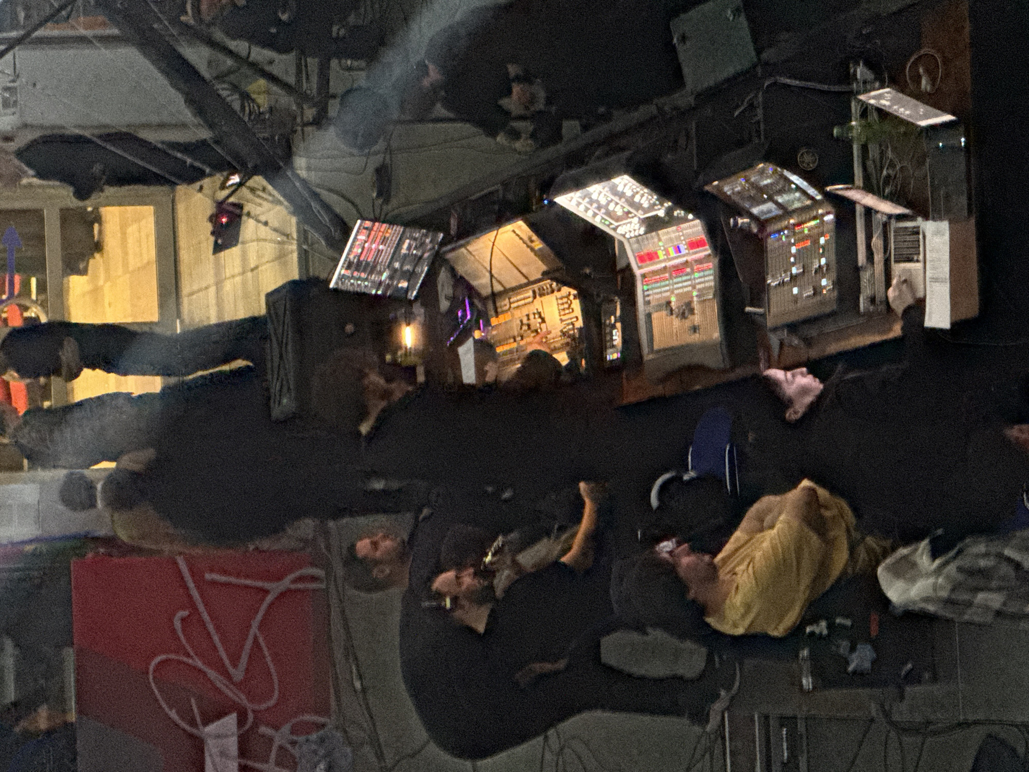 Here's a forensic description of the image:
**Location:** The image appears to be taken inside a building in a city, likely at night. The exact city is unknown. The internal architectural details are not clear from the image.
**Subjects:** The image contains several human figures, all appearing to be adults. Some are standing or sitting, while others are lying down. Their clothing is dark in color, with the exception of one figure wearing a yellow shirt.
**Objects:** Several illuminated control panels with numerous buttons and displays are visible, suggesting a control or monitoring center. There is additional equipment with screens and various buttons. A red object with a white design is also present.
**Actions and Interactions:** Some figures appear to be interacting with the equipment, while others appear to be resting or lying down.
**Environment:** The environment is dimly lit, making it difficult to discern specific details about the room's architecture or furnishings. The building appears to be used for some kind of control or management.