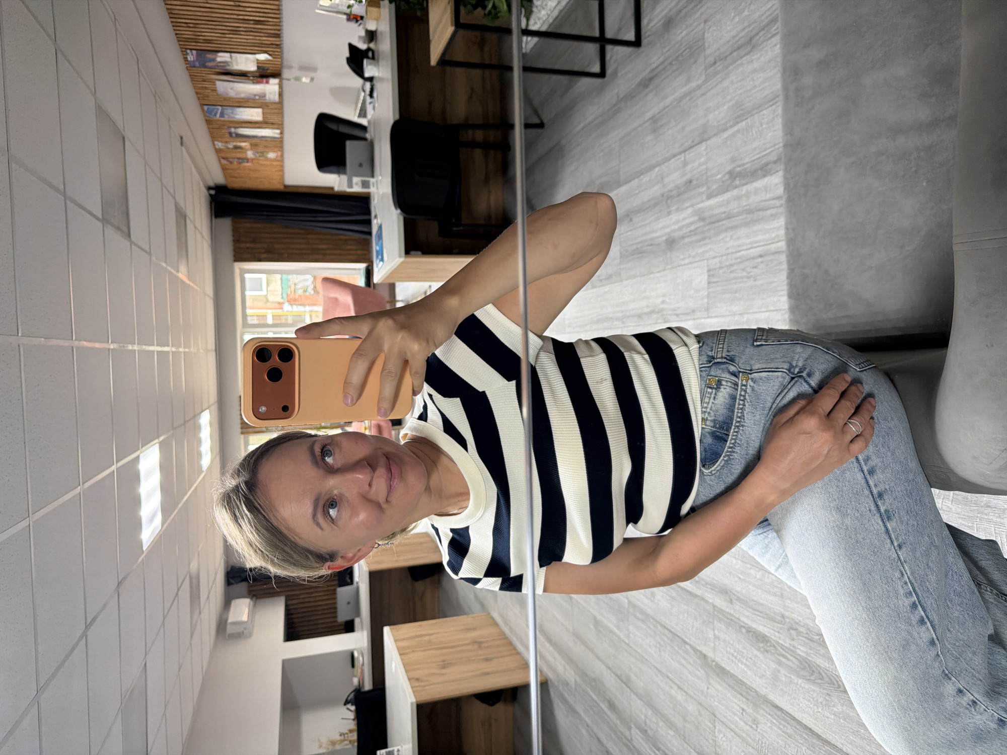 A woman takes a selfie in what appears to be an office space. She is wearing a black and white striped t-shirt and light-wash jeans, and is seated on a gray couch. She holds a peach-colored smartphone up to take the photo, with her left hand partially visible. The background shows office desks, chairs, and a wall with wooden slats and posters. The floor is a light gray wood-grain laminate. The ceiling is a white drop ceiling with fluorescent lighting. The scene is well-lit and appears to be indoors during the daytime. There is no indication of the city of Chaikovsky, Russia.